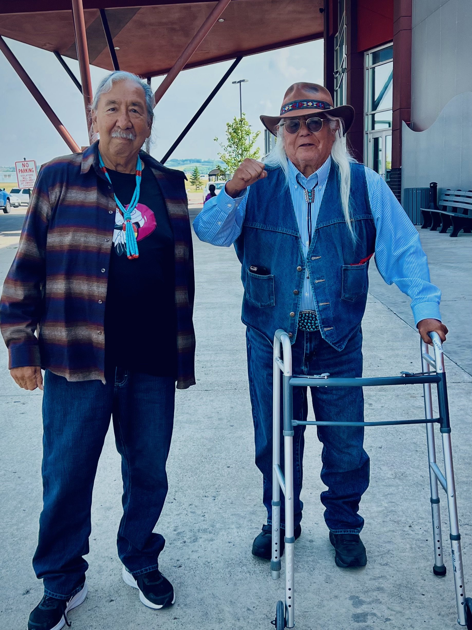 Two elderly men standing outside a building under a covered area; one with a walker's assistance, wearing a vest and sunglasses, the other with long hair and a plaid shirt, smiling with the second man making a fist gesture.