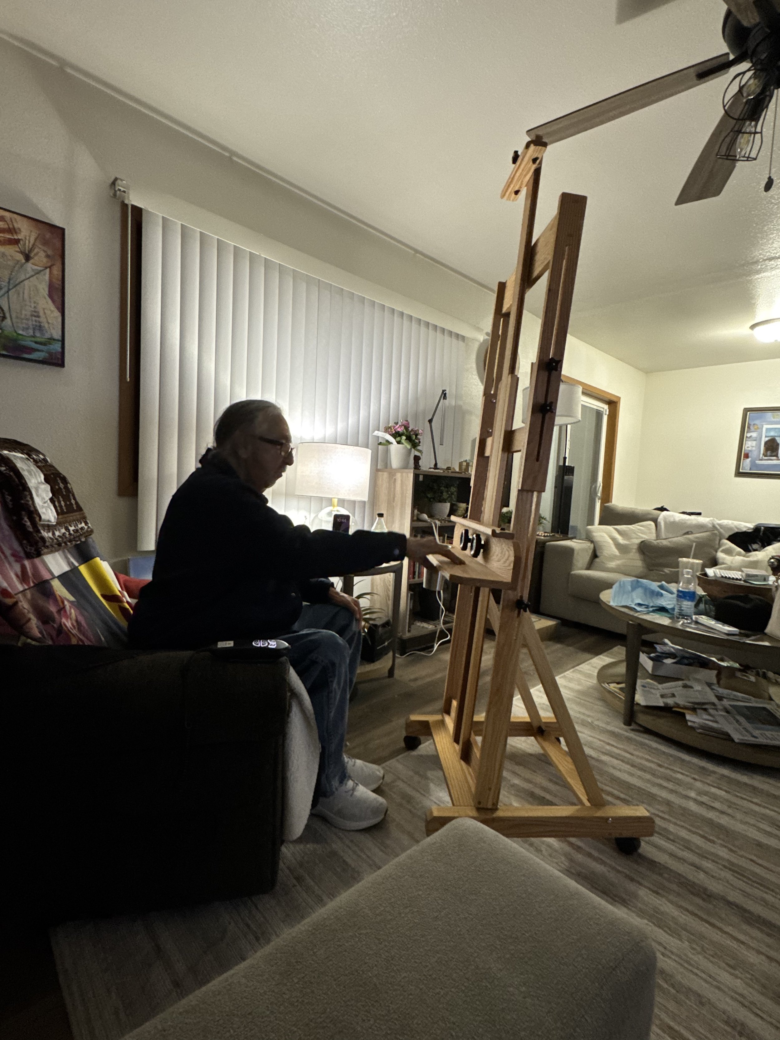 An elderly woman sitting on a dark-colored armchair in a living room, manipulating objects on a large wooden art easel.