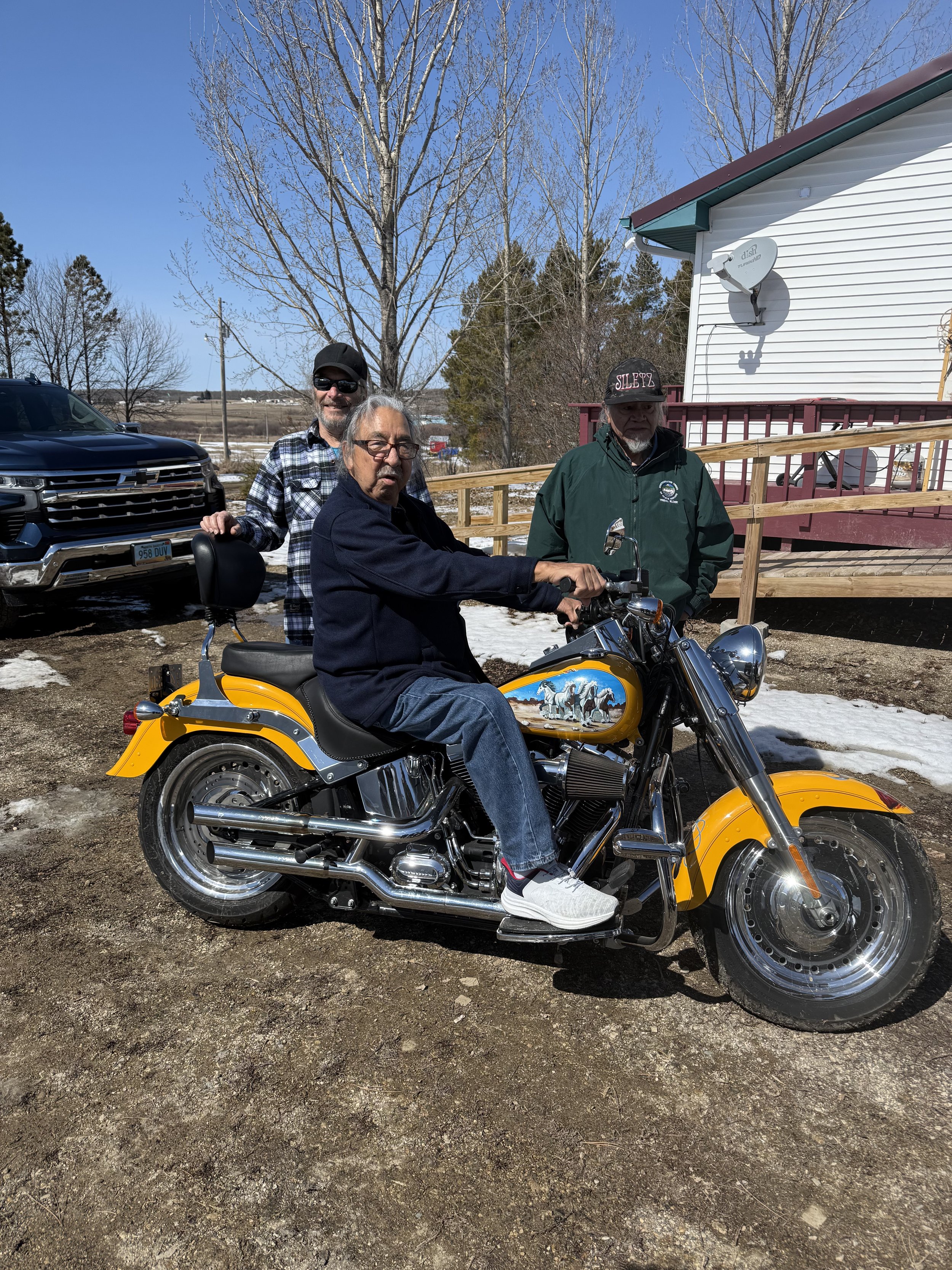 Three men outdoors with a yellow motorcycle, one sitting on it and two standing behind. The setting includes a house with a satellite dish, leafless trees, and patches of snow on the ground, suggesting late winter or early spring. The man on the moto