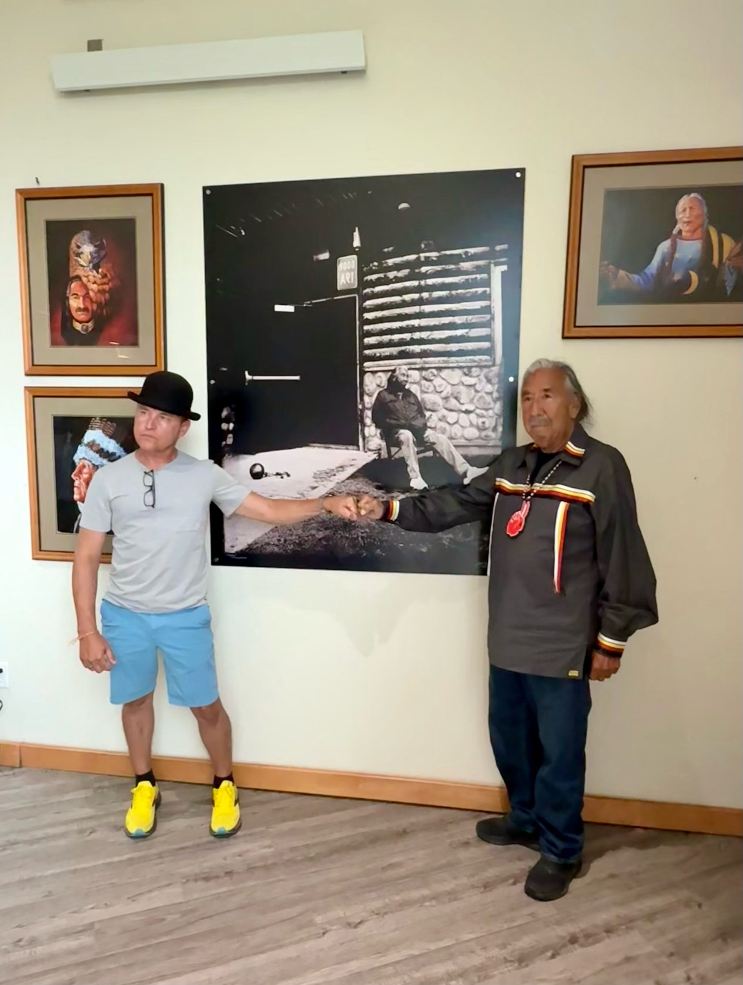 Two men shake hands in front of artwork in a gallery. One man is wearing a gray shirt, blue shorts, yellow sneakers, and a black hat. The other man is dressed in a traditional Indigenous outfit with a medal or decoration. The wall features framed por