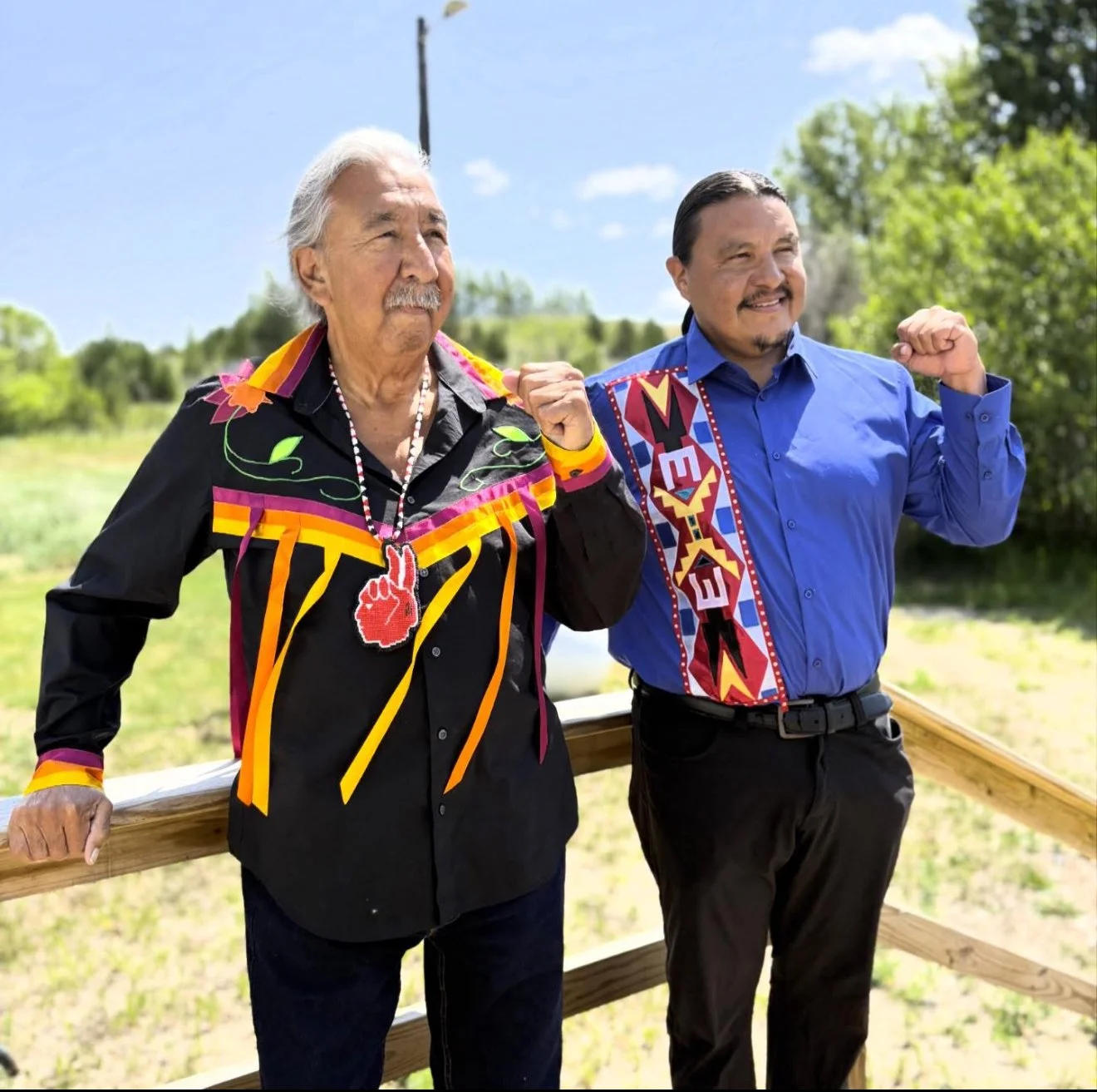 Two men standing outdoors on a wooden railing, flexing their arms, with trees and a blue sky in the background. One man wears a black shirt with colorful designs, and the other wears a blue shirt with a patterned sash.