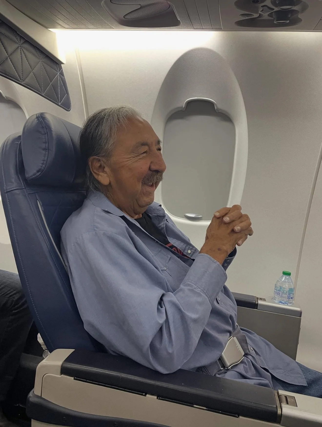 An elderly man with gray hair and a mustache sitting comfortably in an airplane seat, smiling with hands clasped together, next to a window and a water bottle on the tray table.