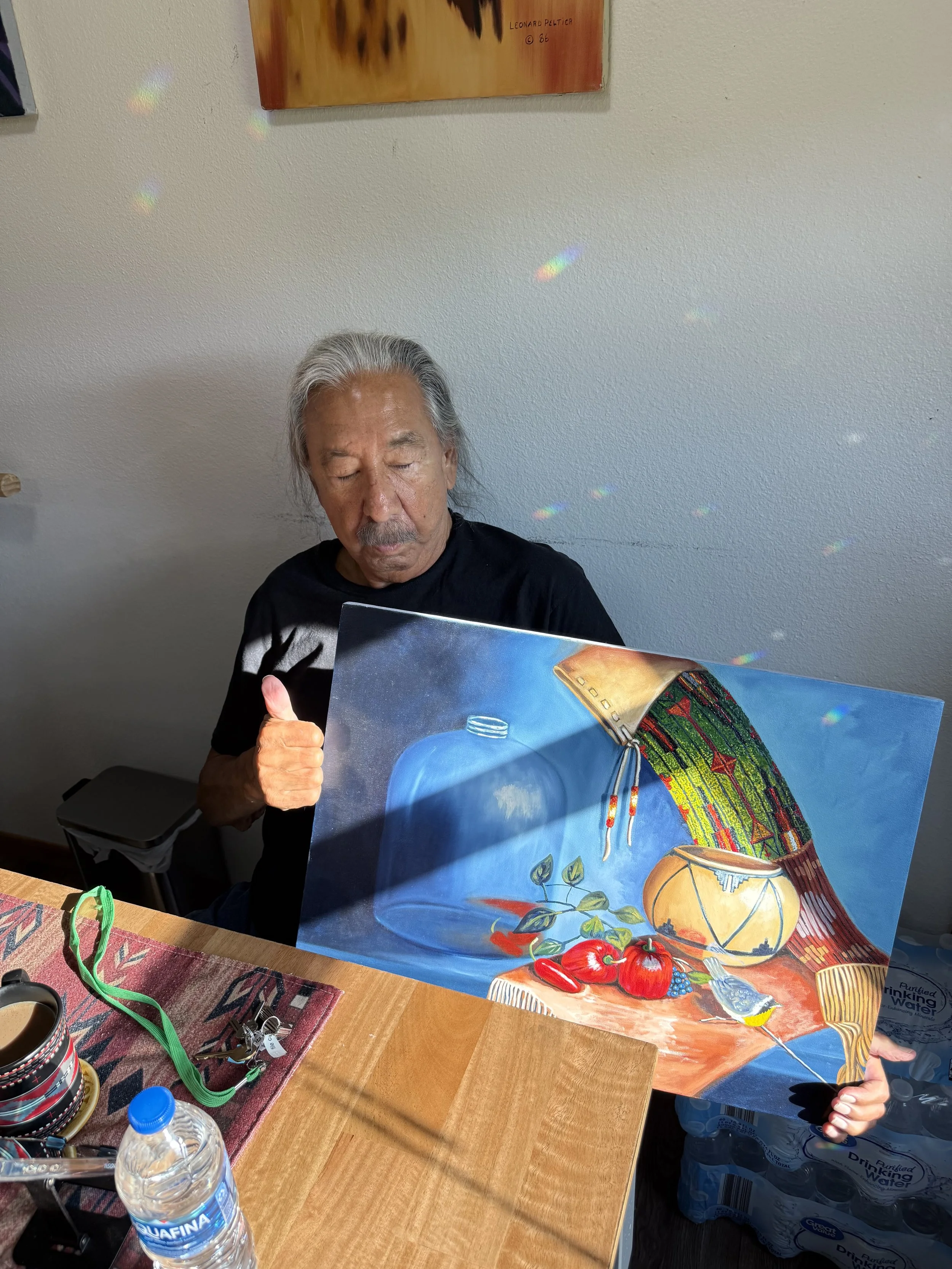 An older man with gray hair giving a thumbs up while holding a colorful still life painting depicting a bottle, a basket with a cloth hanging over it, and various fruits including apples and peppers.