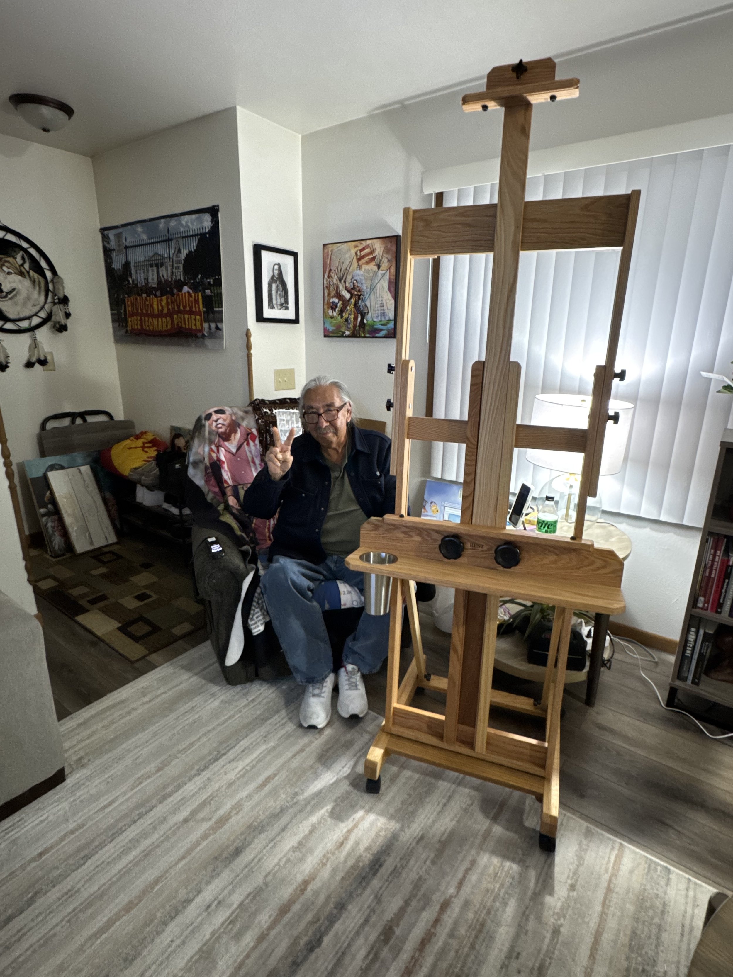 Older man sitting in a living room, making a peace sign, with a large wooden easel in front of him. The room has framed artwork on the walls, a couch with colorful pillows, and a table with a lamp and other items.