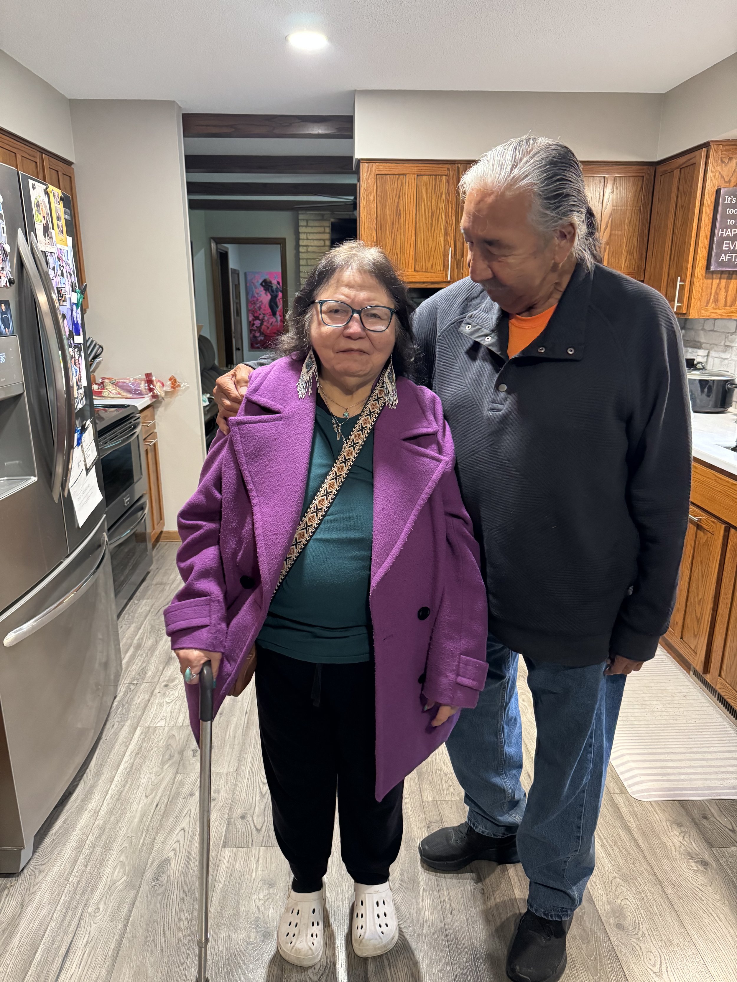 An elderly woman with glasses, wearing a purple coat, holding a cane, standing in a kitchen with a man who has long gray hair, wearing a black jacket, both smiling softly and standing close together.