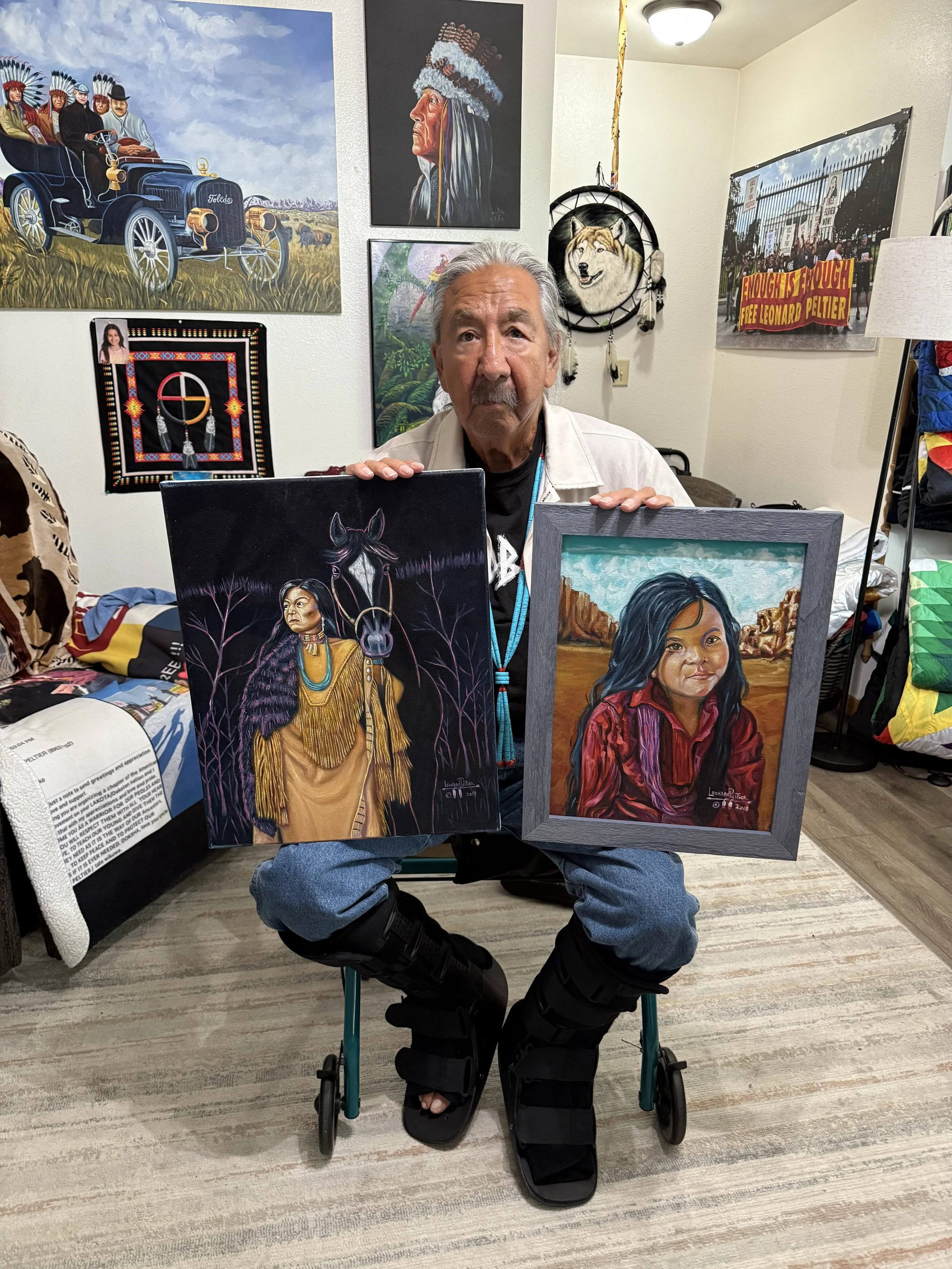An elderly man with gray hair and a mustache holding two framed paintings inside an art studio. The painting on the left depicts a Native American woman with long hair, traditional clothing, and a horse, set against a dark background with trees. The