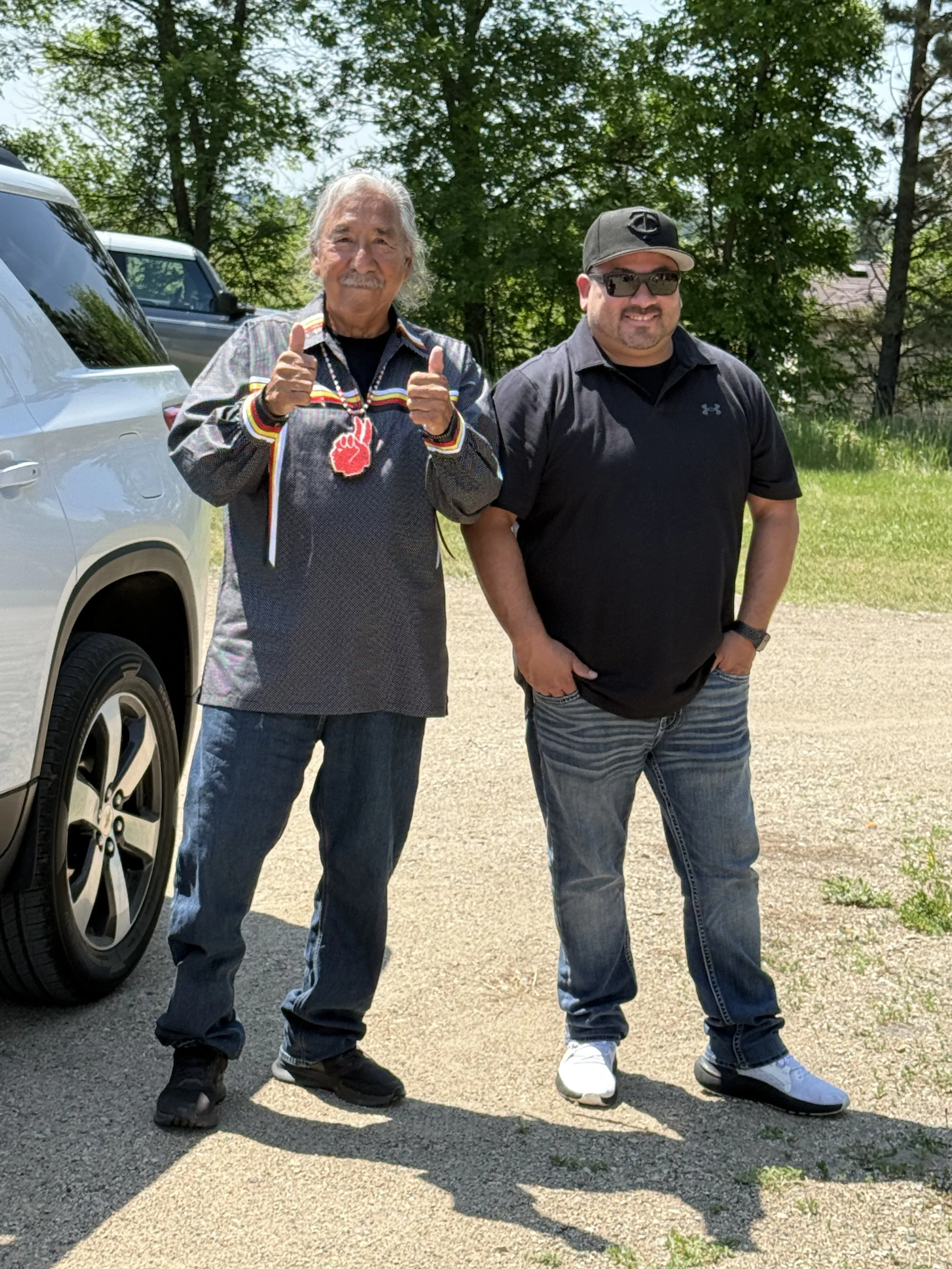 Two men standing outdoors on a sunny day, smiling, with trees in the background. One man is older, wearing a gray shirt with colorful stripes and a necklace, giving a thumbs-up. The other man is younger, dressed in a black t-shirt, jeans, sunglasses, and a cap, with his hands in his pockets.