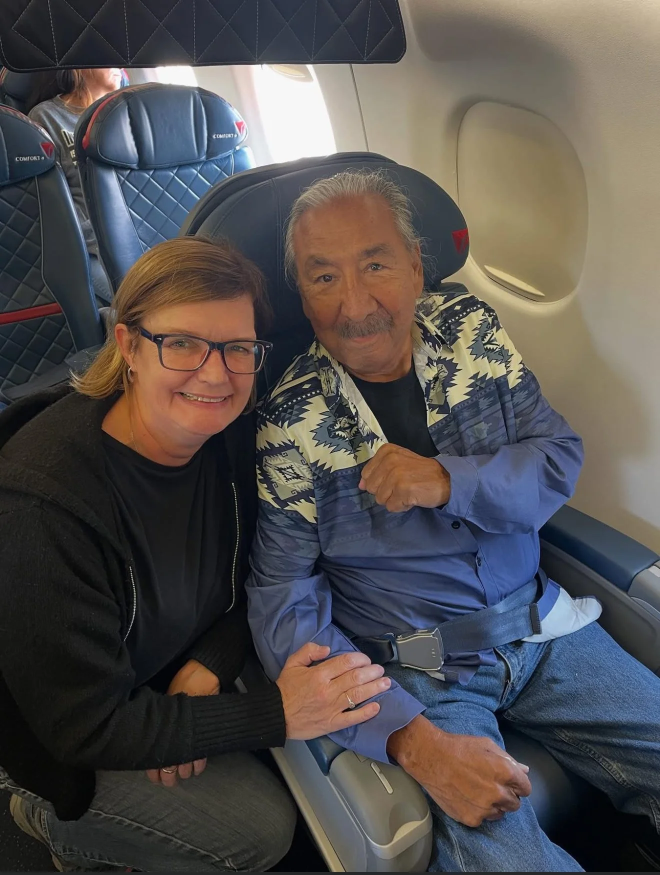 A woman and an older man seated next to each other on an airplane. The woman has short brown hair, glasses, and is smiling. The man has gray hair, a mustache, and is wearing a patterned shirt and jeans. They appear happy and are posing for the photo