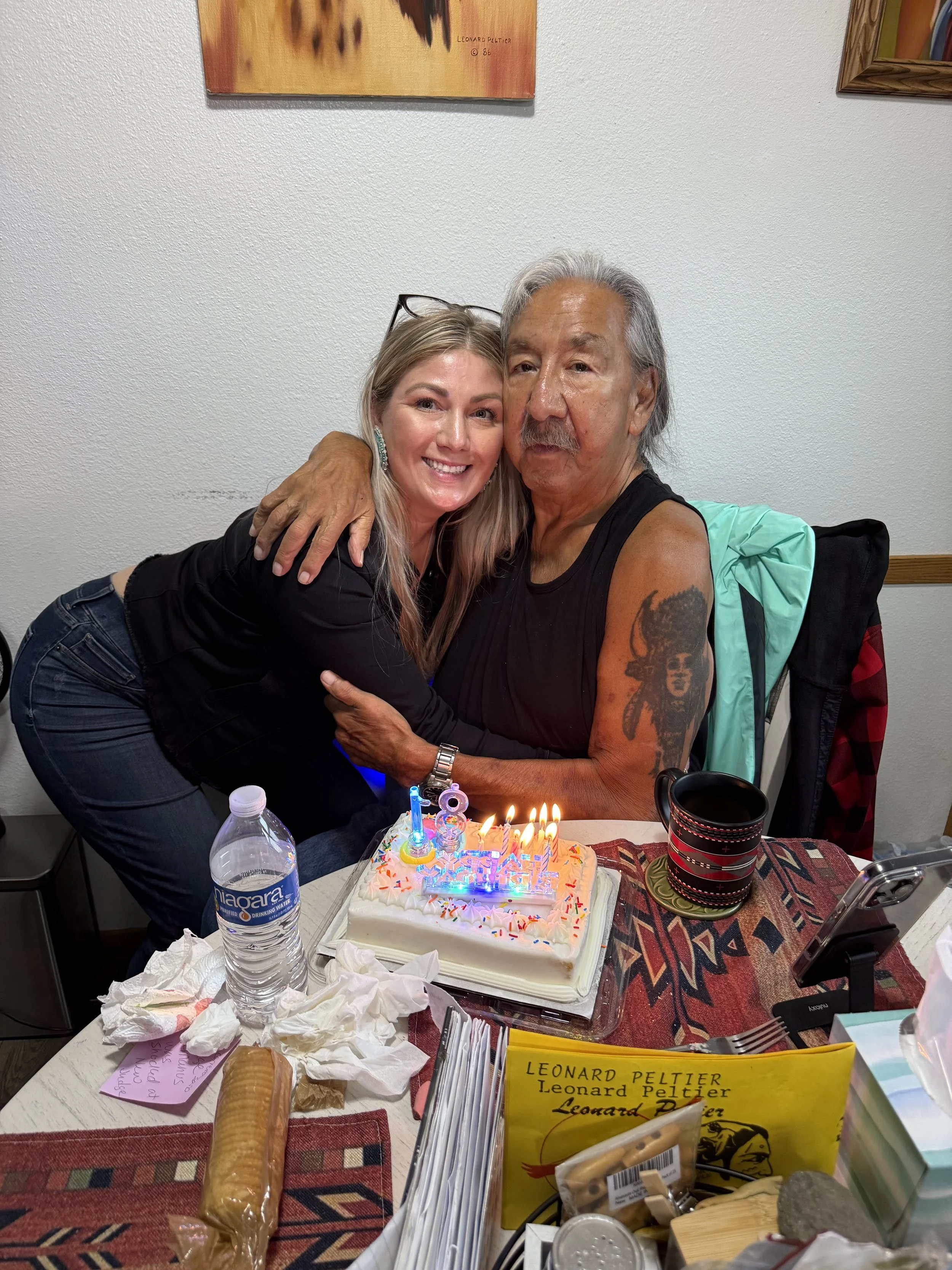 A woman and an older man are hugging and smiling at a birthday celebration. The man is sitting in a chair with a birthday cake with candles in front of him, and the woman is leaning over the table, both appearing happy. The table has a water bottle,