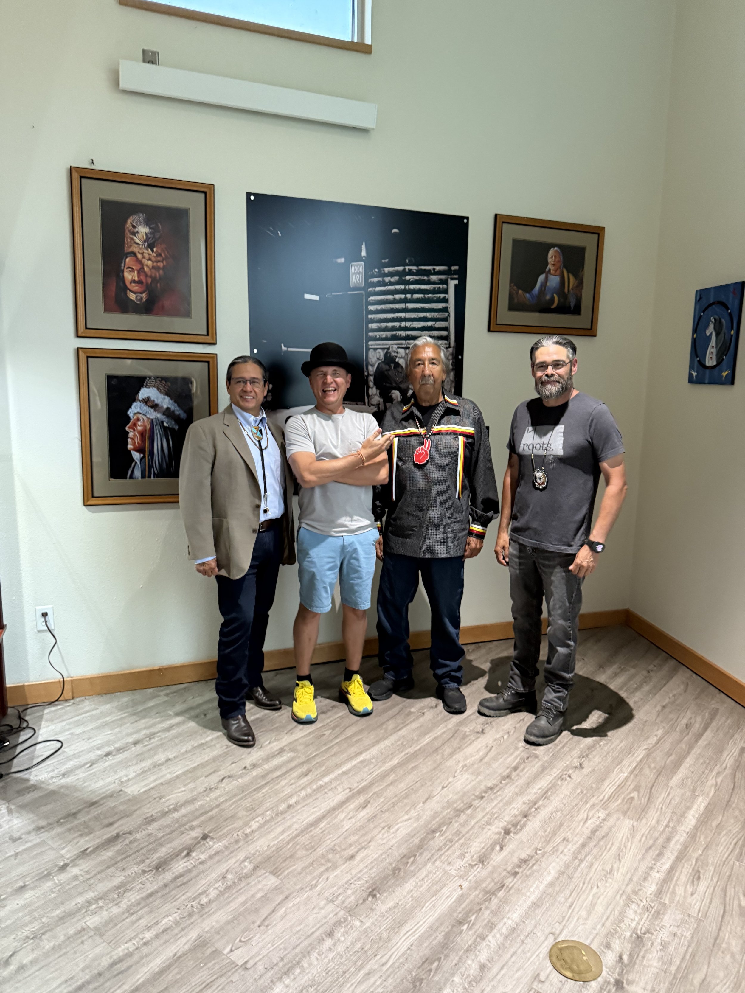 Four men standing together inside an art gallery, smiling at the camera. Behind them are four framed pieces of art and a large black-and-white photograph. The man second from the left is wearing a hat and casual clothes, while the others wear more fo