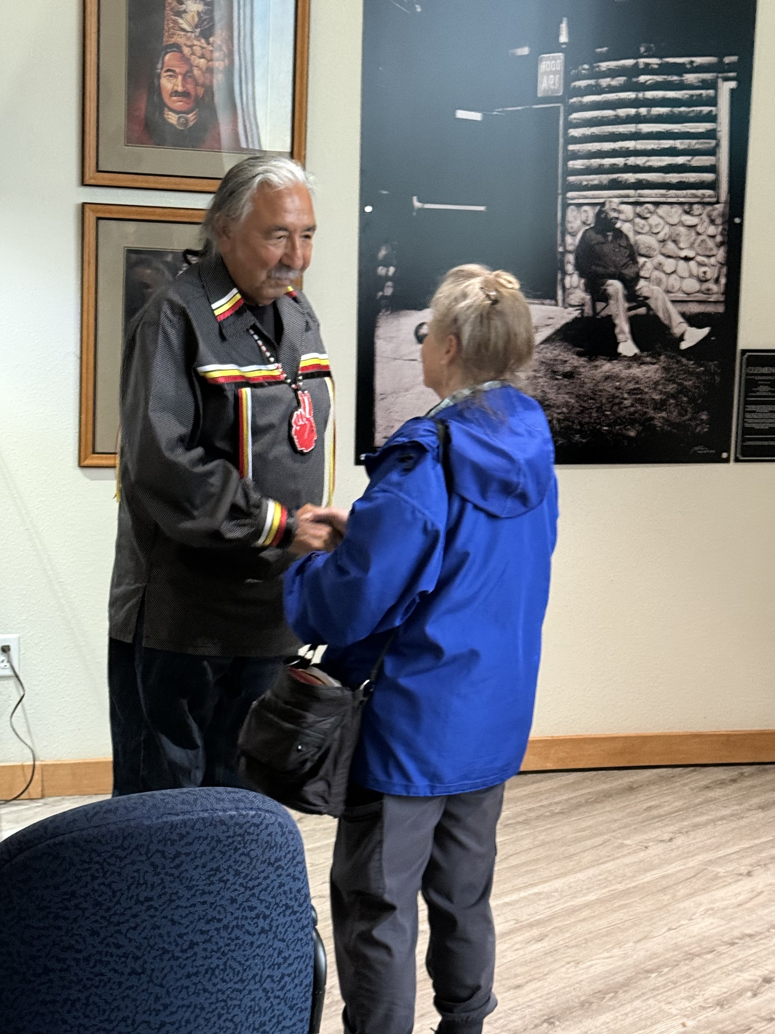 An elderly man and woman are shaking hands inside a room with framed photos on the wall. The man is wearing a black shirt with traditional Indigenous patterns and a necklace with a medallion. The woman is wearing a blue jacket and glasses. There is a