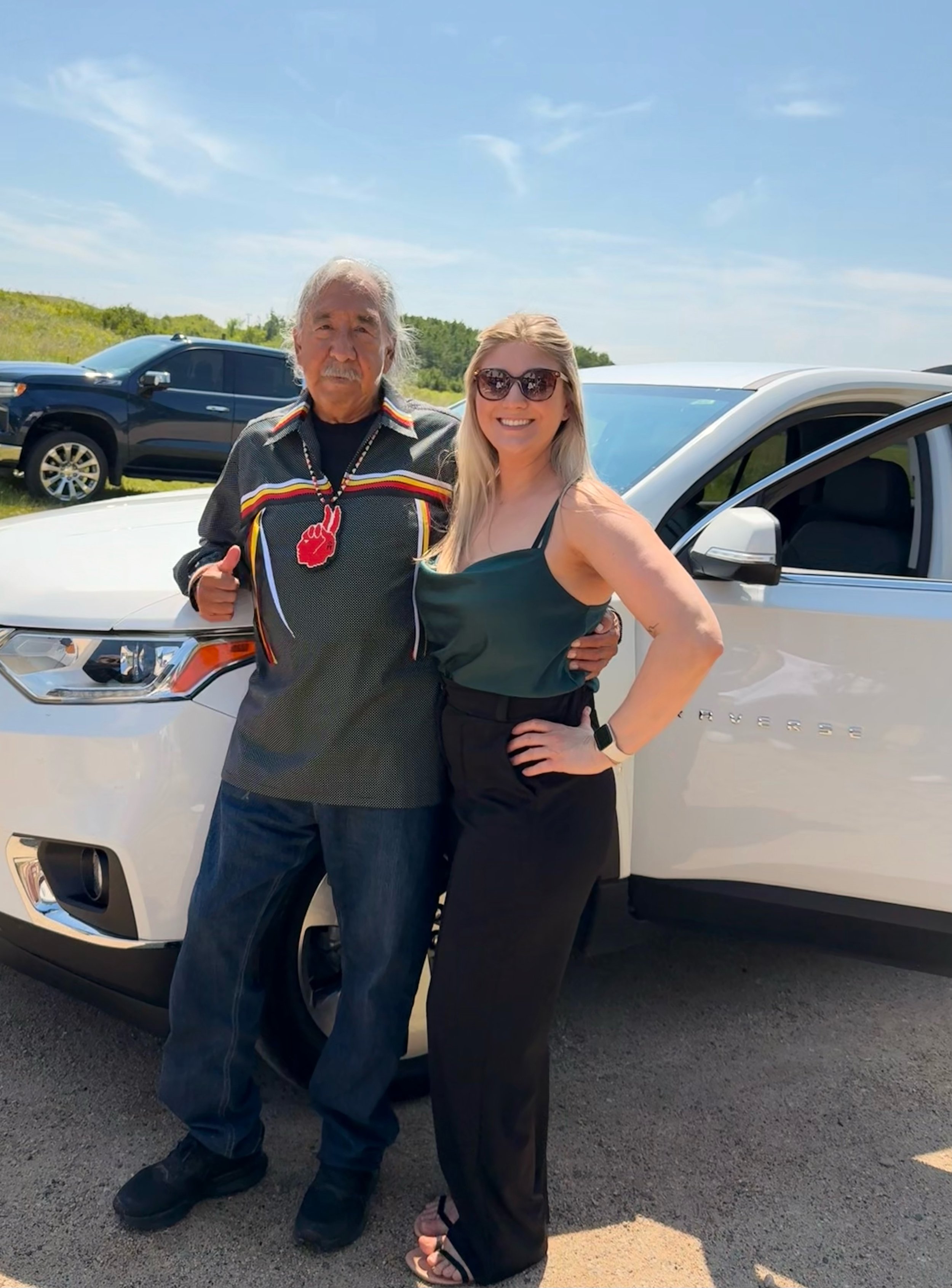 A man and a woman standing together outdoors near a white Chevrolet Traverse, smiling at the camera. The man has gray hair, a mustache, and is wearing a colorful shirt with a red, yellow, and black pattern, with a red beaded necklace. The woman has l