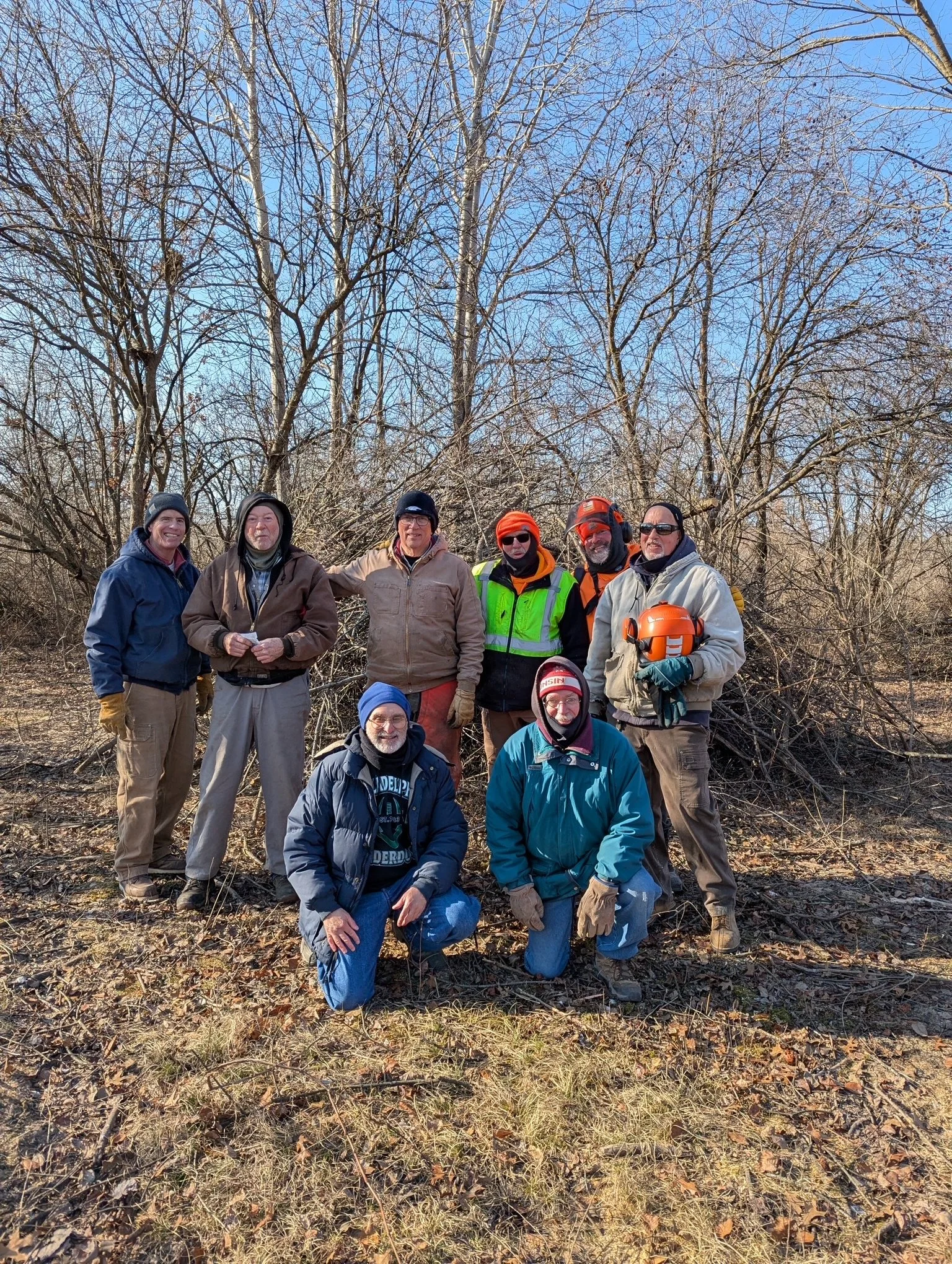 Group of nine adults dressed in winter clothing standing outdoors in a leafless wooded area on a sunny day.