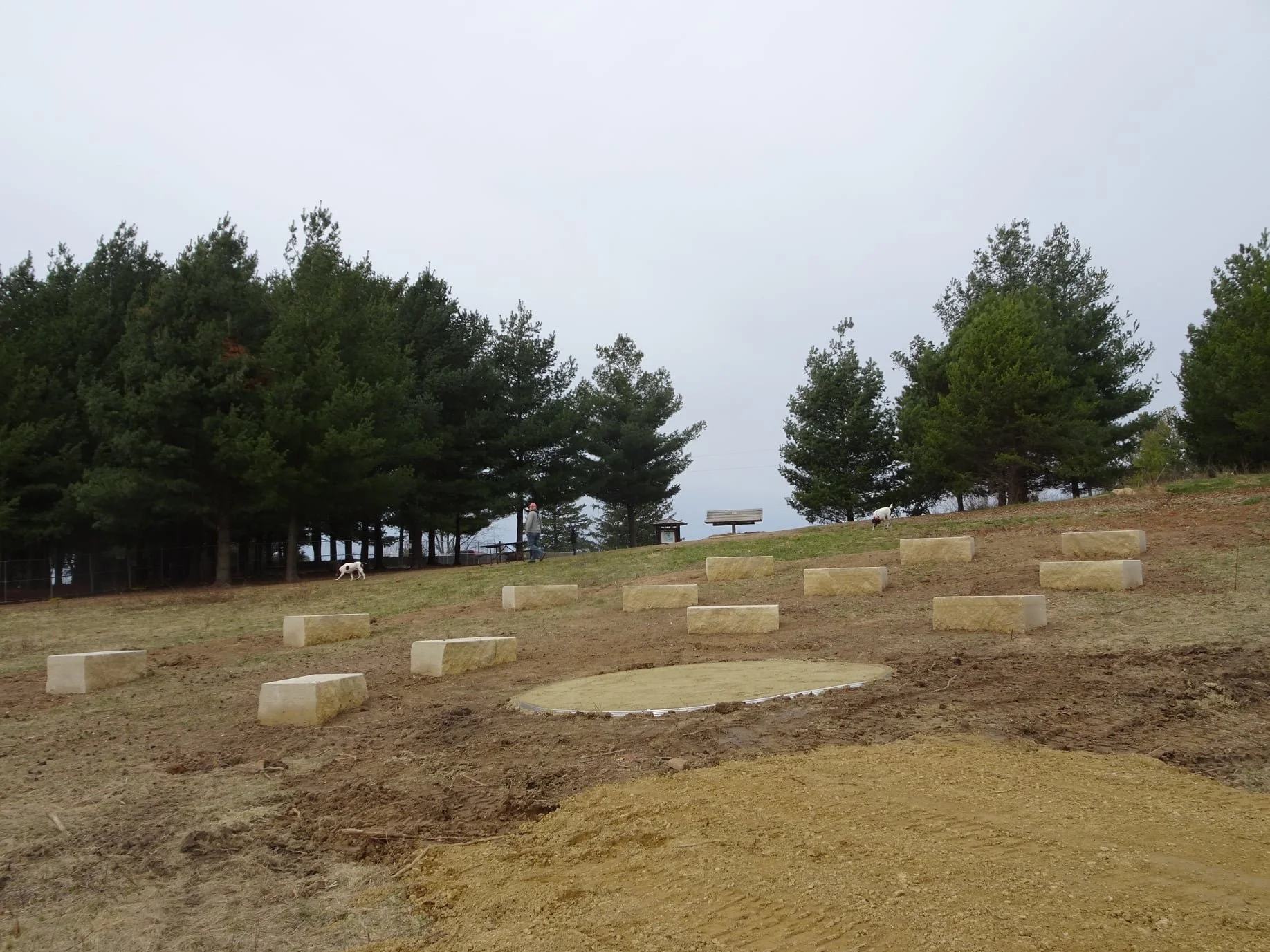 Bare dirt area with concrete blocks arranged in elevation steps, two benches, a grassy hill, with trees and a person walking dogs in the background.