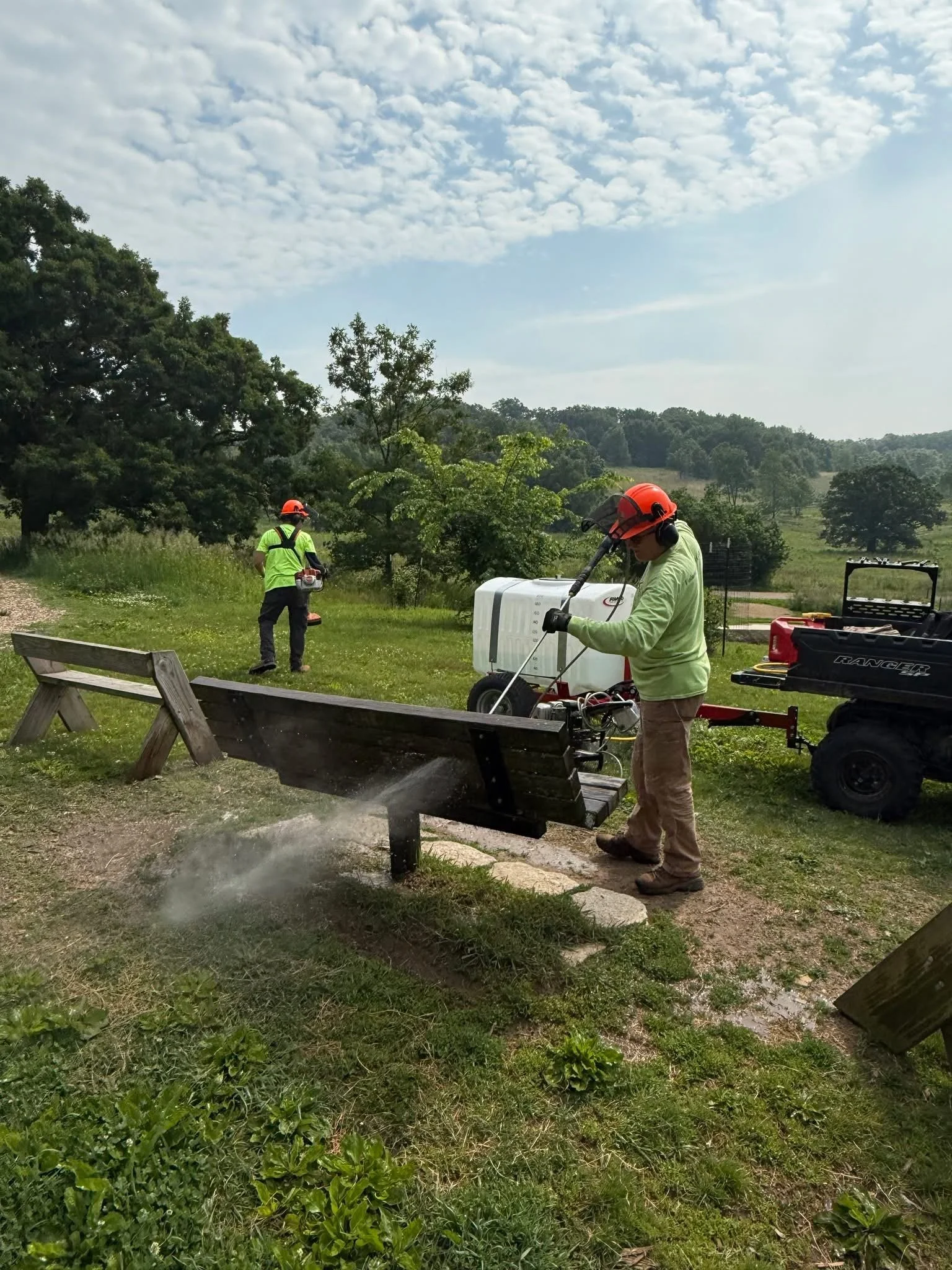 Two workers wearing safety helmets and ear protection are cleaning a park bench with a high-pressure washer in a green outdoor setting with trees and grass.