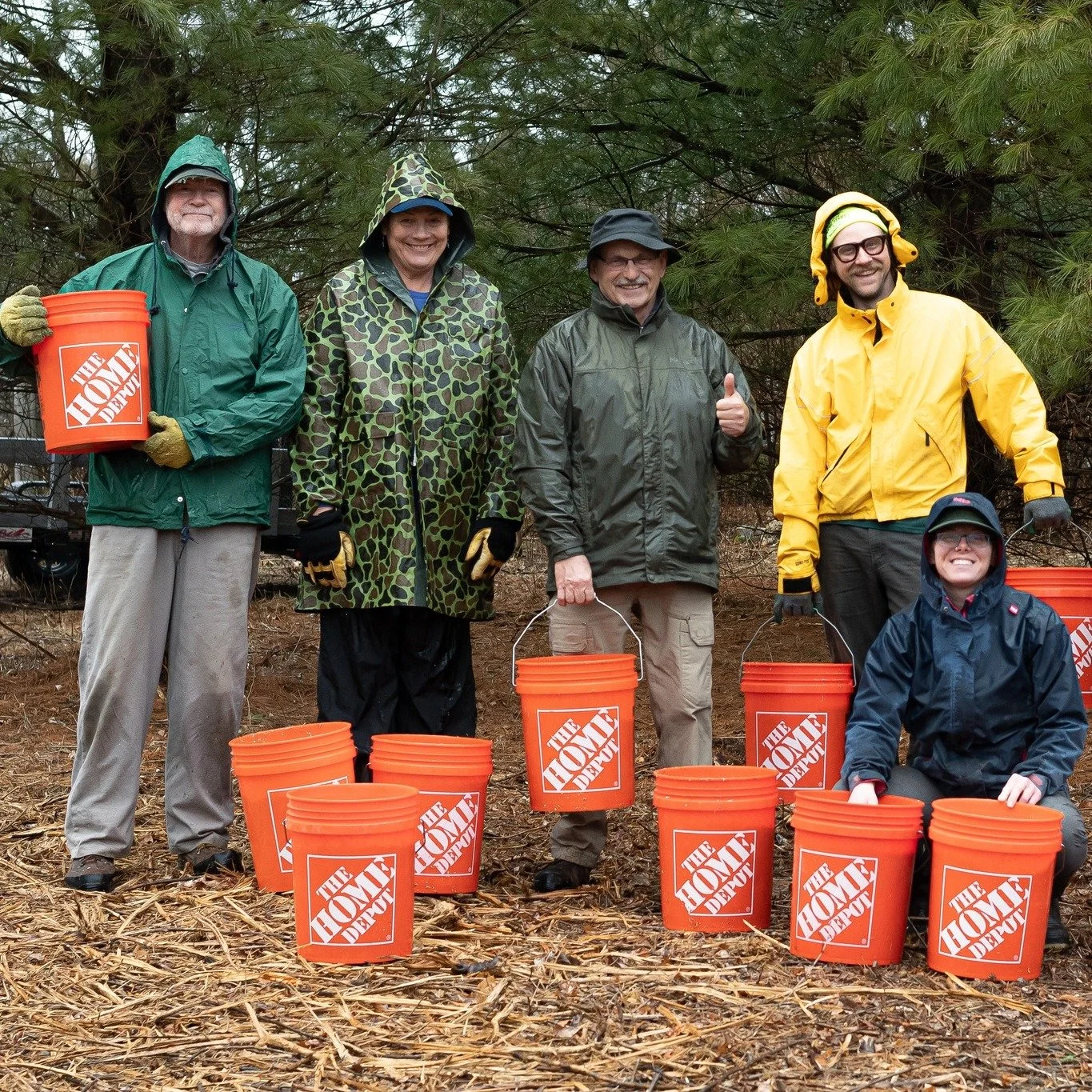 Group of six people dressed in rain gear holding orange buckets with The Home Depot logo outdoors in a wooded area.