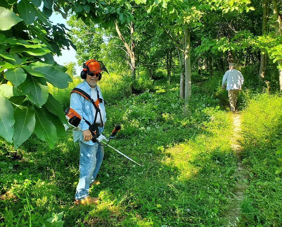 A man wearing safety gear, including an orange helmet and ear protection, using a string trimmer in a lush green forest area. Another person is seen walking away on a clearing path.
