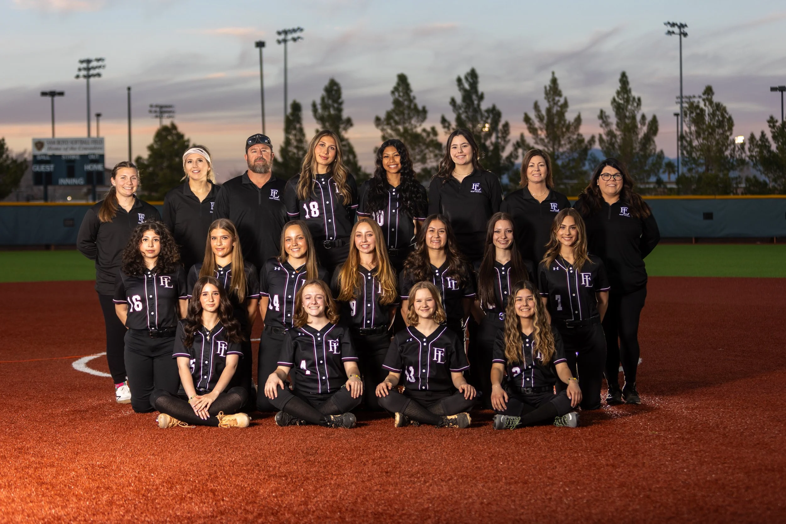 A softball team photo on a field during evening sunset, featuring players in black uniforms and coaches in black jackets, with some team members sitting and others standing.