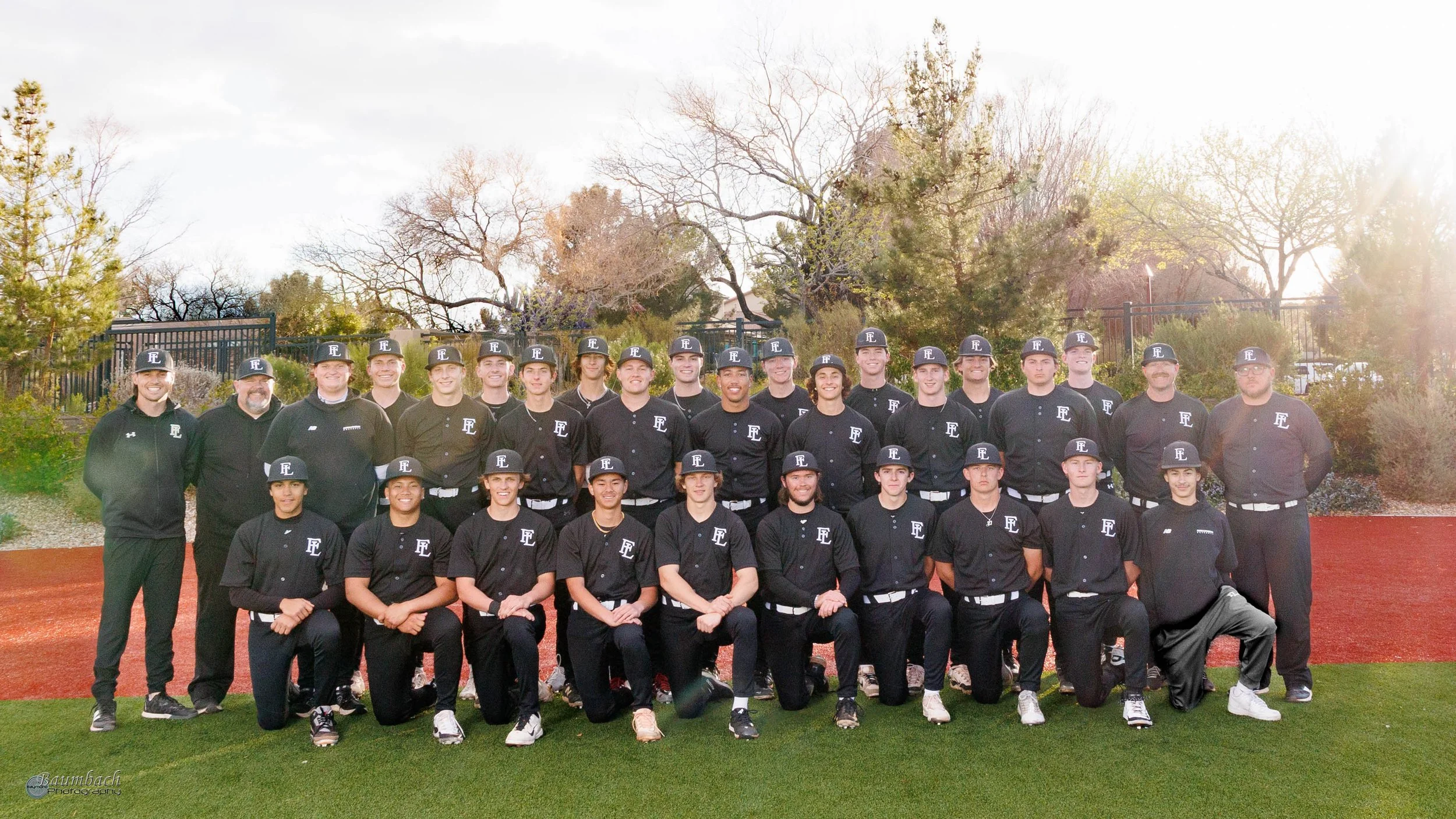 A baseball team pose for a group photo outdoors on a sunny day. They are dressed in black uniforms with 'EL' logos on caps and shirts, standing and kneeling on a field with a red dirt area and green grass. Behind them are trees with sparse leaves and a fence.