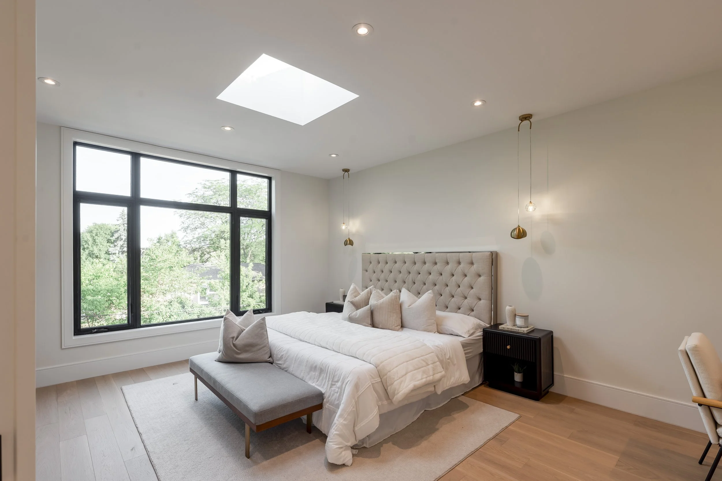 Modern bedroom with large window overlooking green trees, beige upholstered bed with multiple pillows, black nightstands, pendant lights, and a skylight in the ceiling.