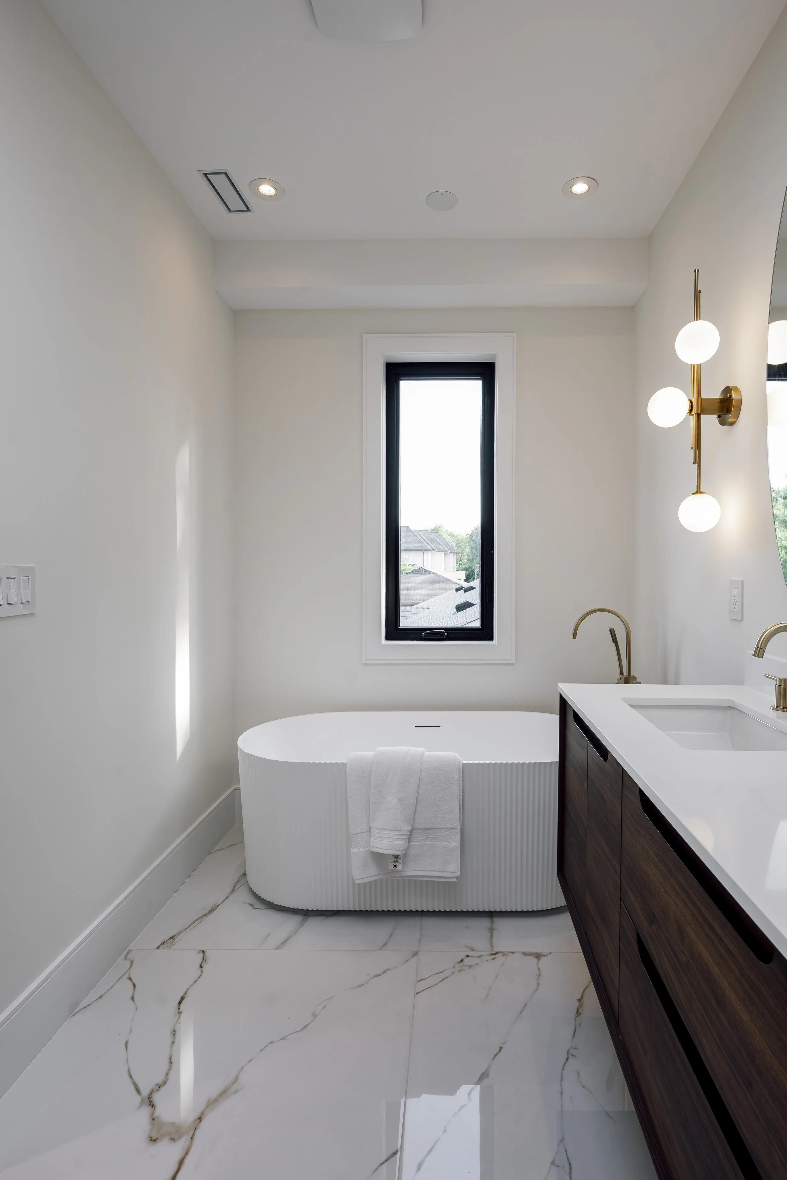 Modern bathroom with a white bathtub, dark wood vanity, white countertop, glass window, and golden light fixtures.