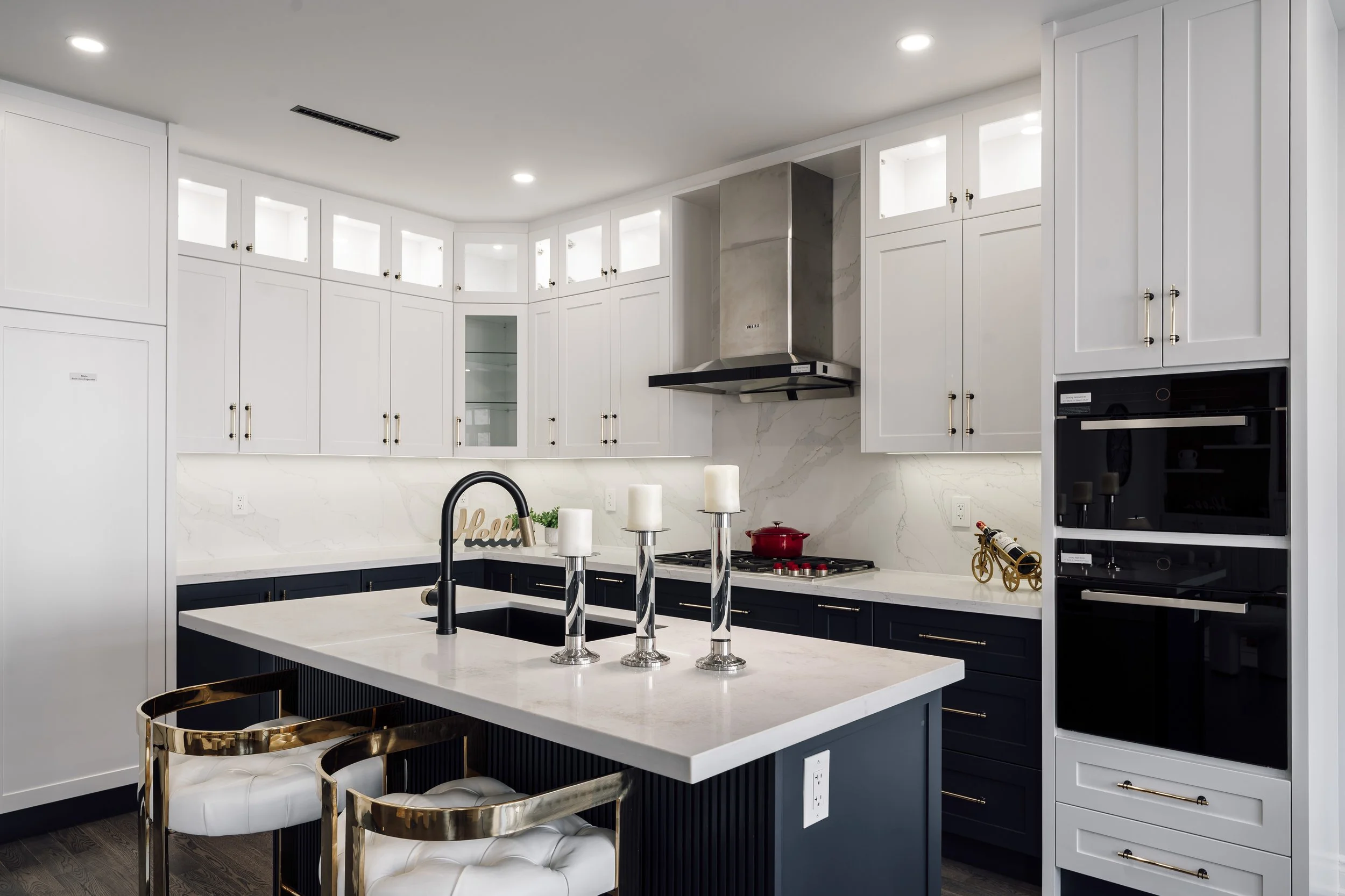 Modern kitchen with white upper cabinets, dark lower cabinets, a central island with a white countertop, a black faucet, and brass accents. The kitchen has built-in appliances, a stainless steel range hood, and decorative candles on the island.