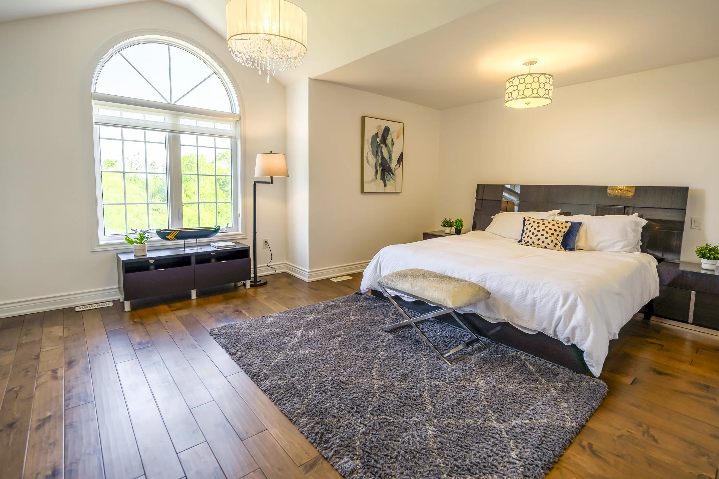 A bedroom with a large bed, dark wood headboard, white bedding, and decorative pillows. A bench at the foot of the bed. Two matching nightstands with plants. A large window with an arched top, a black console table, a potted plant, and a boat sculptu