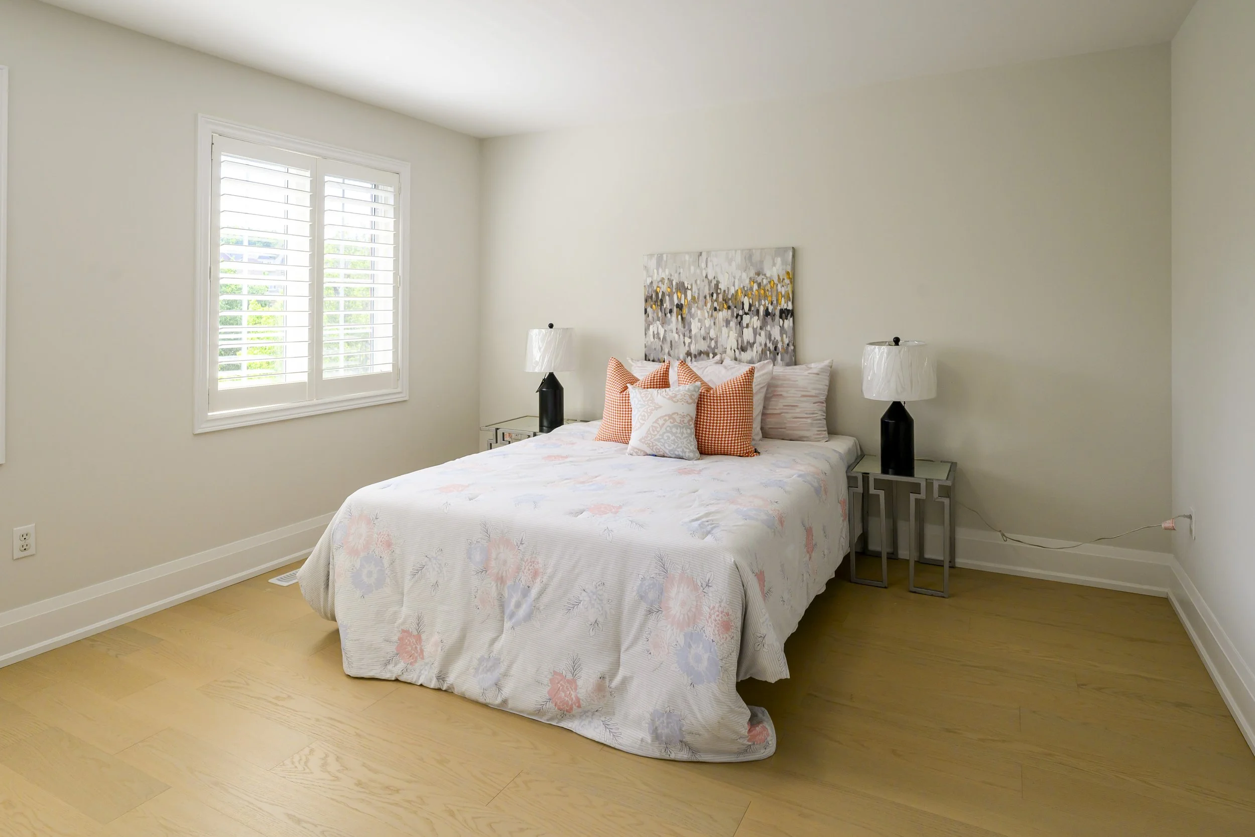 Bedroom with a bed, floral bedding, orange and white pillows, artwork above the headboard, two nightstands with black lamps, window with white shutters, light wood flooring, cream painted walls.