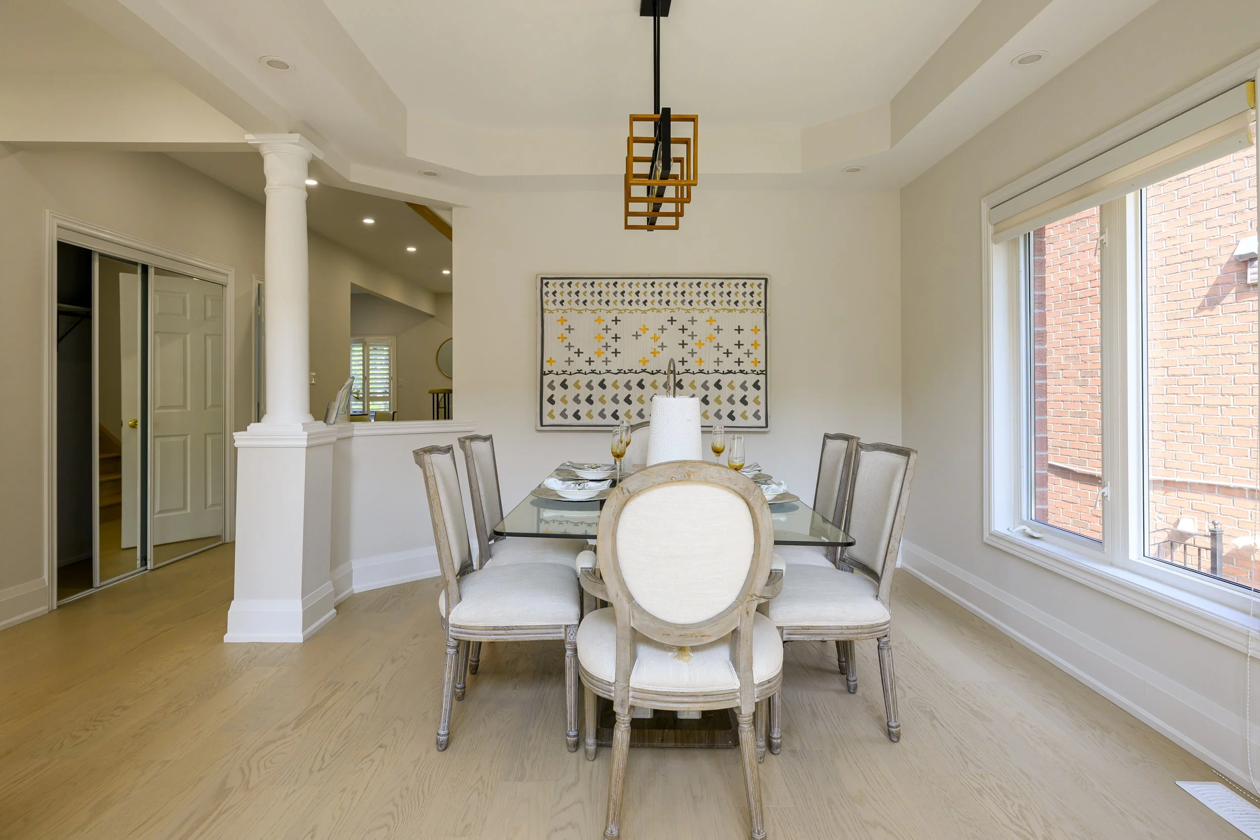 Dining room with a glass table, six chairs, a large window with a brick building outside, and a modern geometric wall art piece