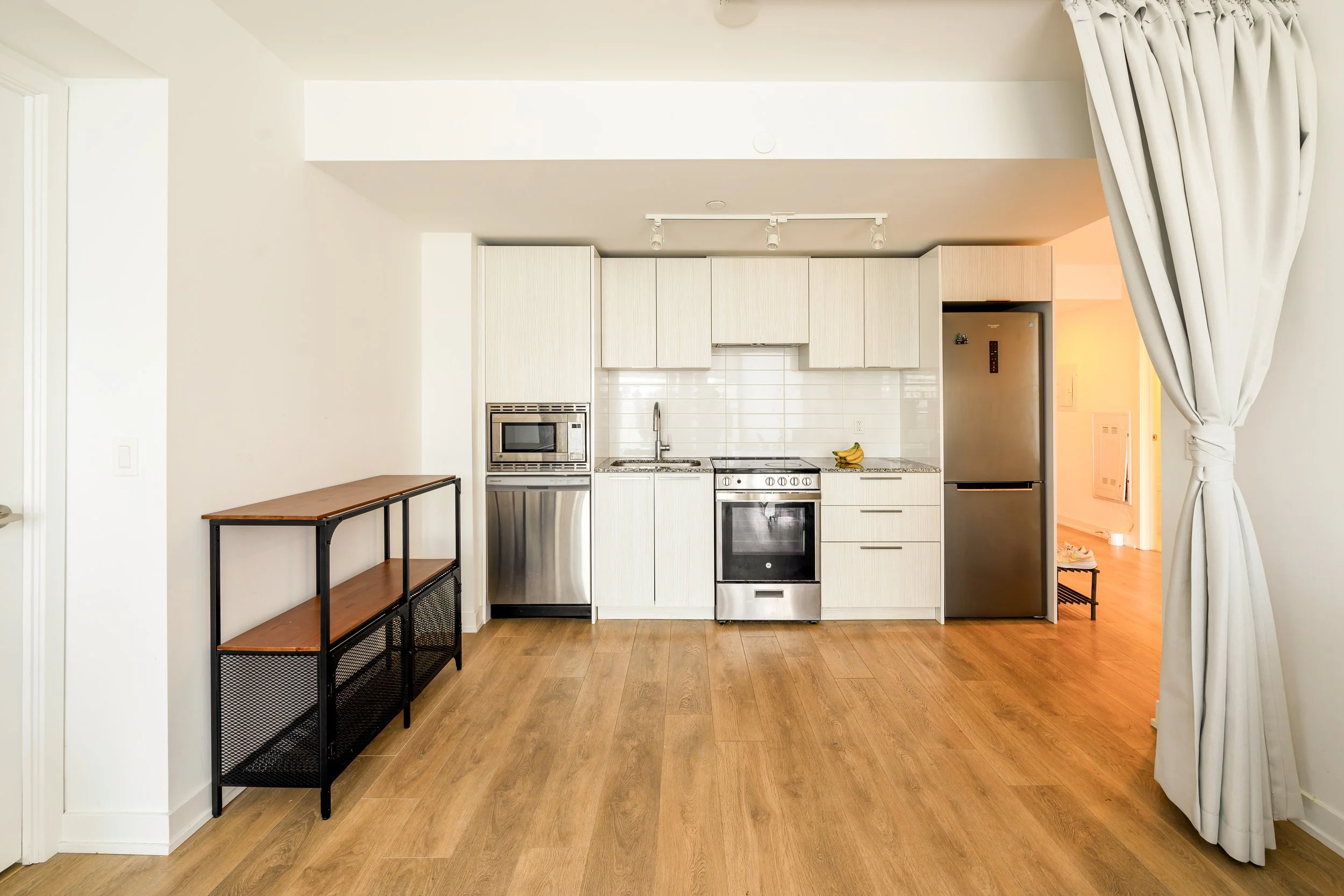 Empty modern kitchen with white cabinets, stainless steel appliances, and wooden flooring. There is a narrow black and wood shelving unit on the left and a large window with curtains on the right.