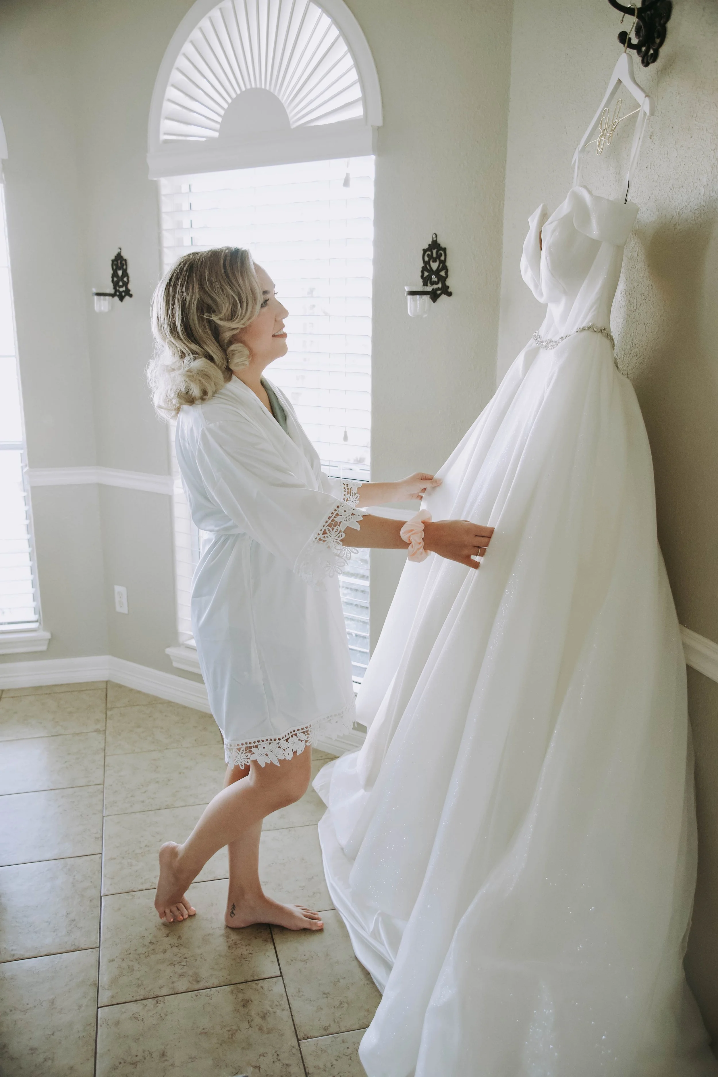 A woman in a white lace-trimmed robe is looking at a white wedding dress hanging on a hook on the wall. She is smiling and holding the dress with both hands, standing on a tiled floor near a window with blinds.
