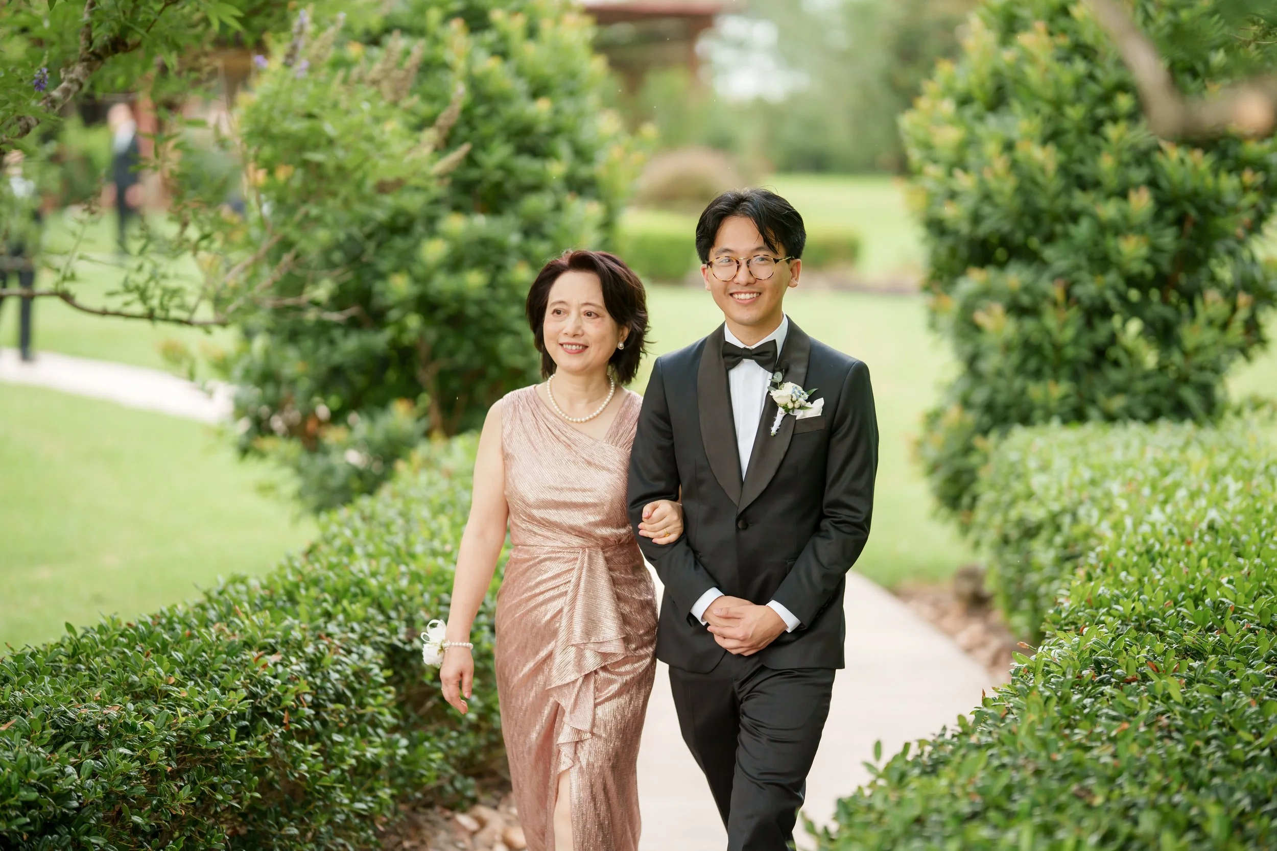 A young man in a tuxedo walking arm-in-arm with an older woman in a formal dress through a garden pathway.