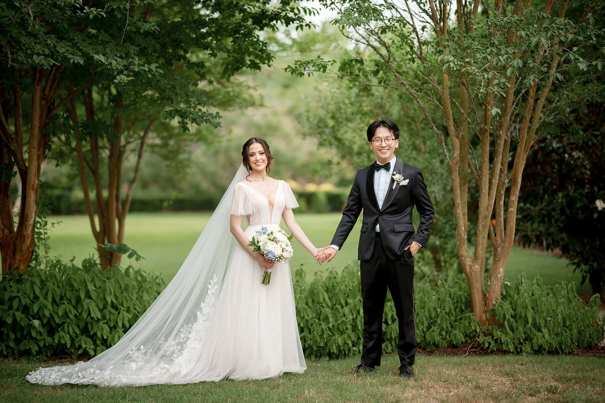 A bride and groom standing outdoors in a garden, holding hands and smiling. The bride is wearing a white wedding gown with a long veil and holding a bouquet of flowers. The groom is wearing a black tuxedo with a bow tie, glasses, and a boutonniere.