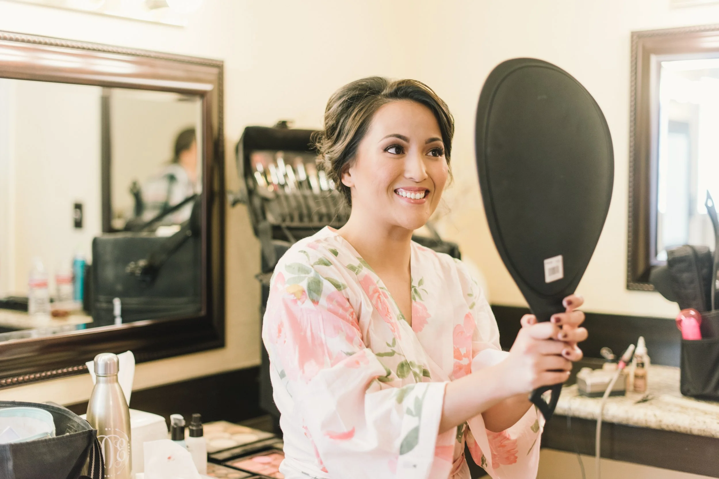 A woman in a floral robe holding a black hand mirror in a makeup room, smiling at her reflection.