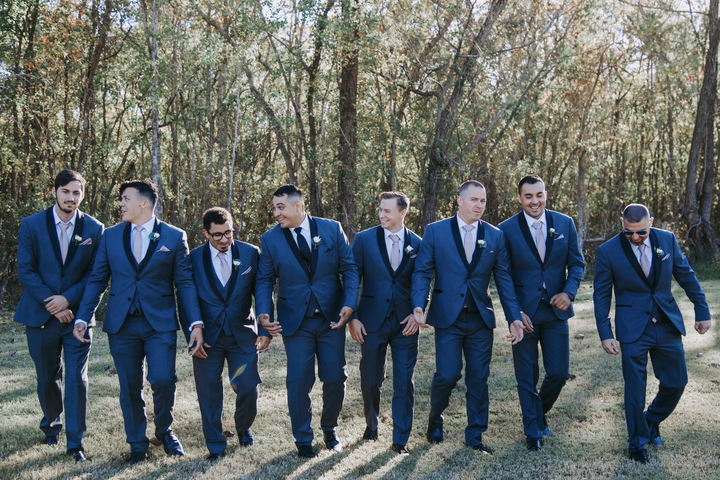 A group of nine men in matching blue suits walking together outdoors in a grassy area with trees in the background.