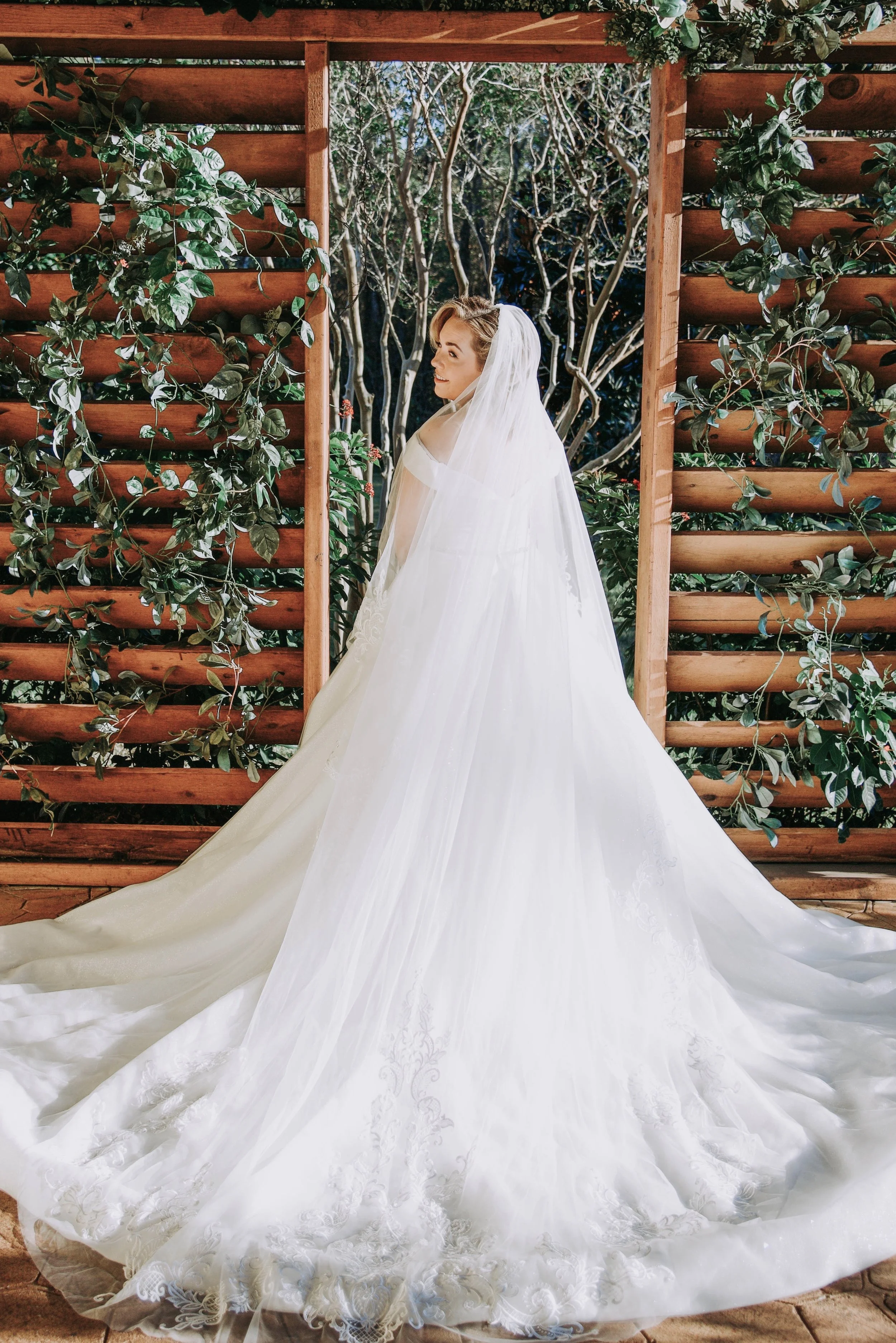 A bride in a white wedding gown with a long train and veil, standing in front of a wooden structure with green foliage and trees in the background.