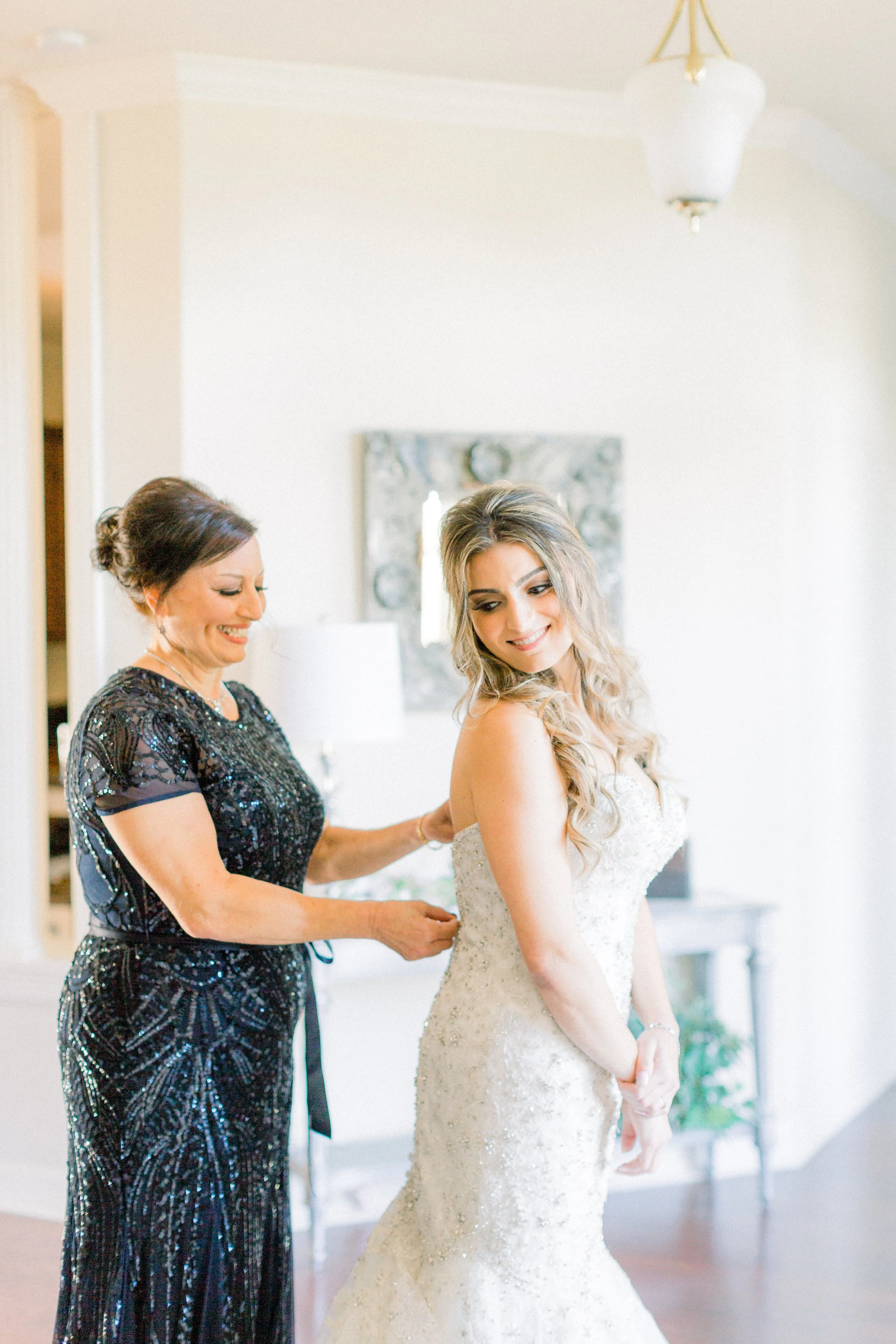 A bride in a white wedding gown getting help with her dress from a woman in a black sequined dress in a well-lit room.