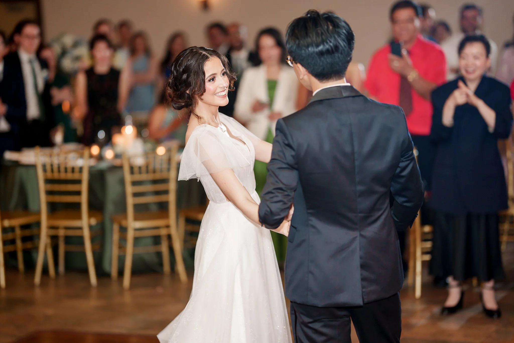 A bride and groom sharing a dance at their wedding reception with guests watching and smiling in the background.