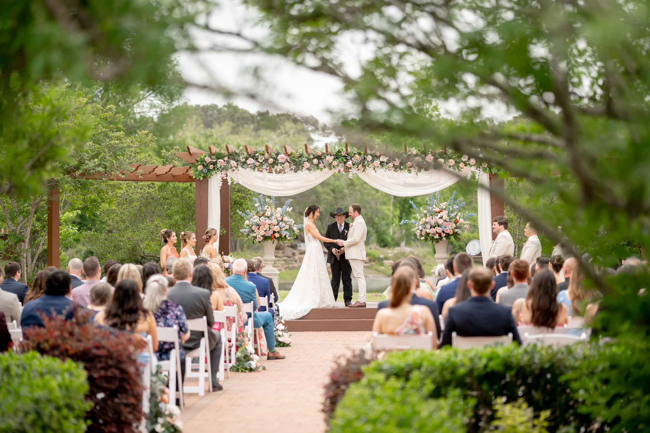 A wedding ceremony outdoors with a bride and groom exchanging vows, surrounded by guests, with floral arrangements and a wooden arch decorated with drapery and flowers.