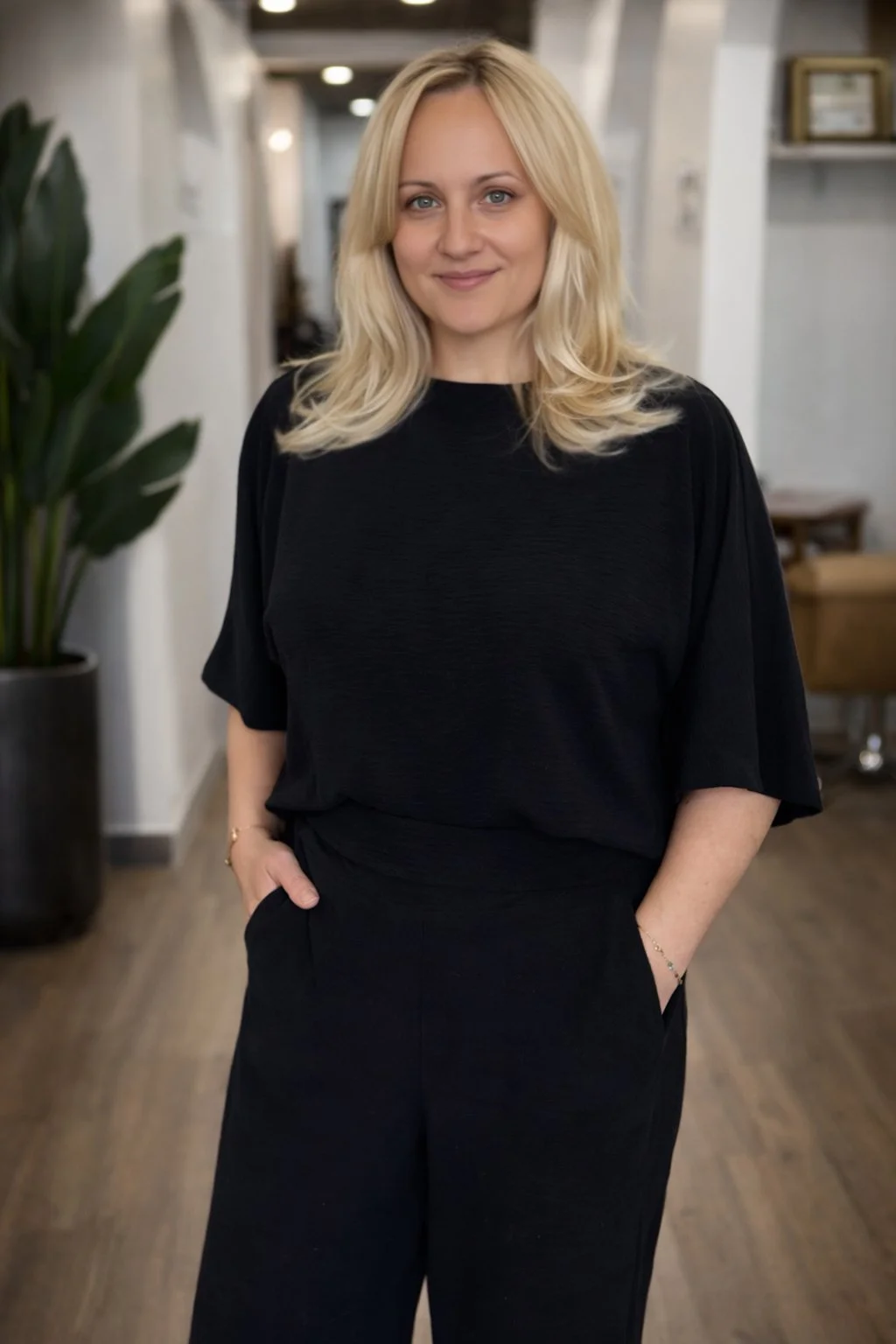 A woman with blonde hair wearing a black top and black pants, standing indoors with a large green plant on her left side and a blurred background.