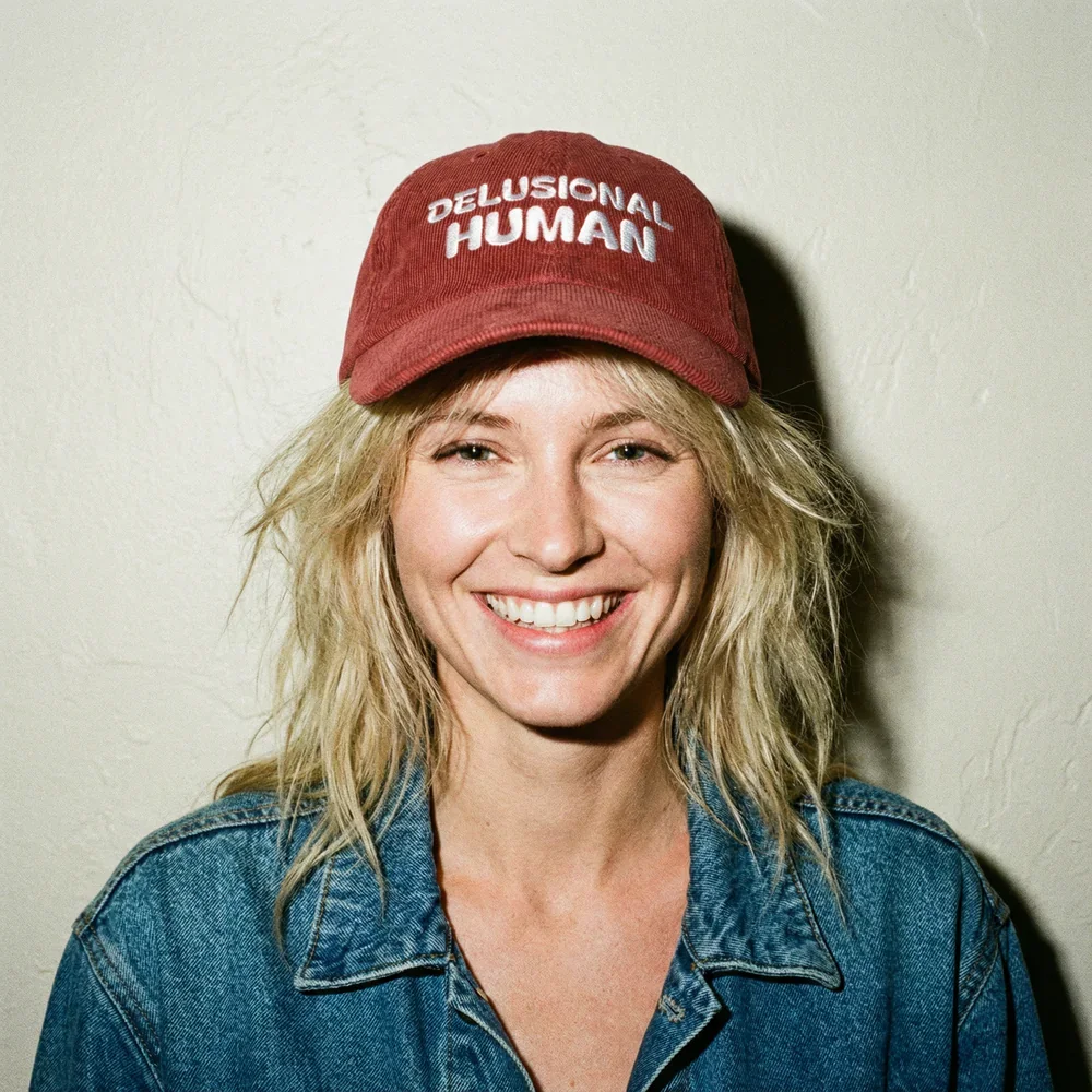 A woman with blonde hair smiling, wearing a red cap that says 'Delusional Human' and a denim jacket, standing against a plain wall.