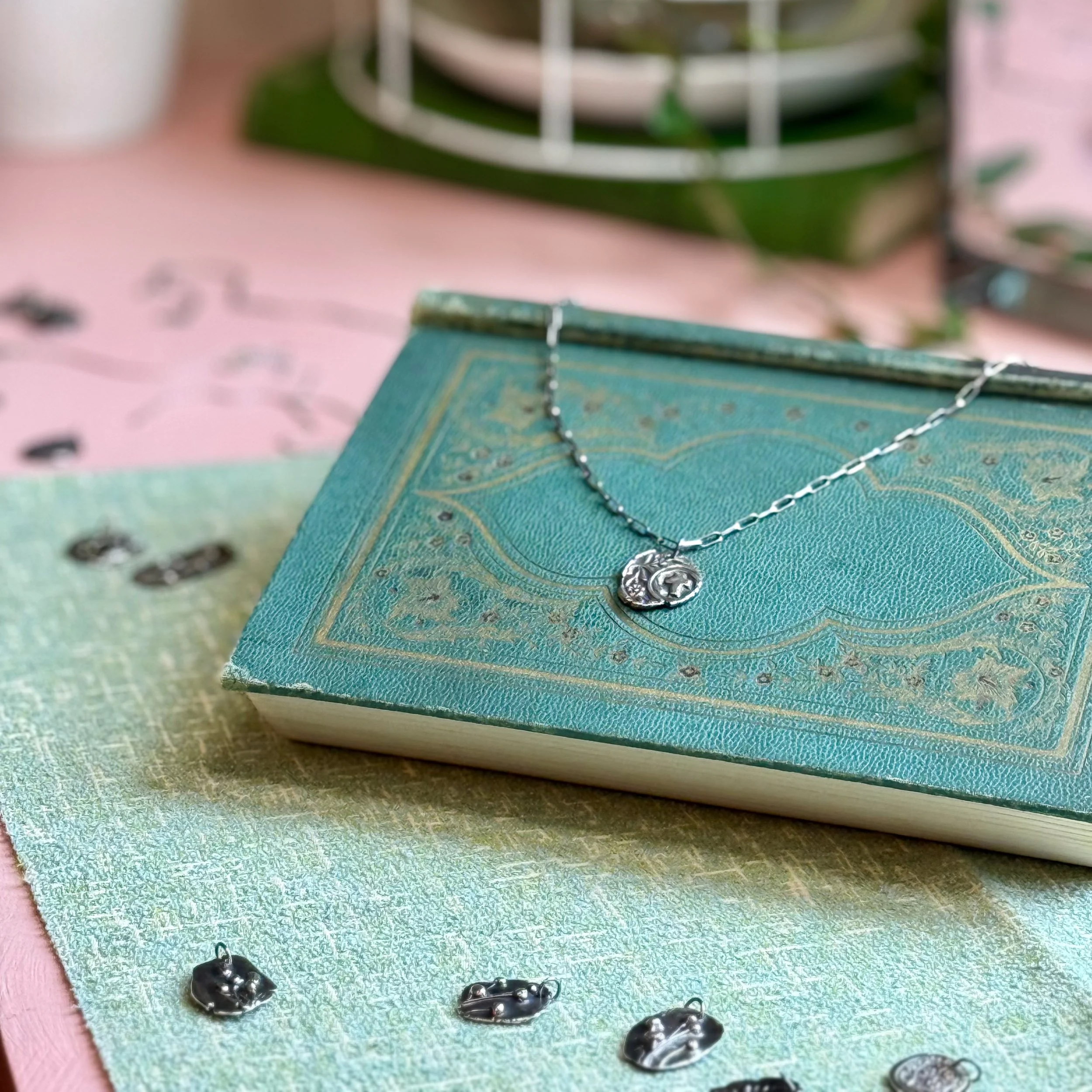 A silver necklace with a circular pendant resting on a vintage turquoise hardcover book. The book is placed on a pink surface with scattered small metallic necklace charms and an out-of-focus background.