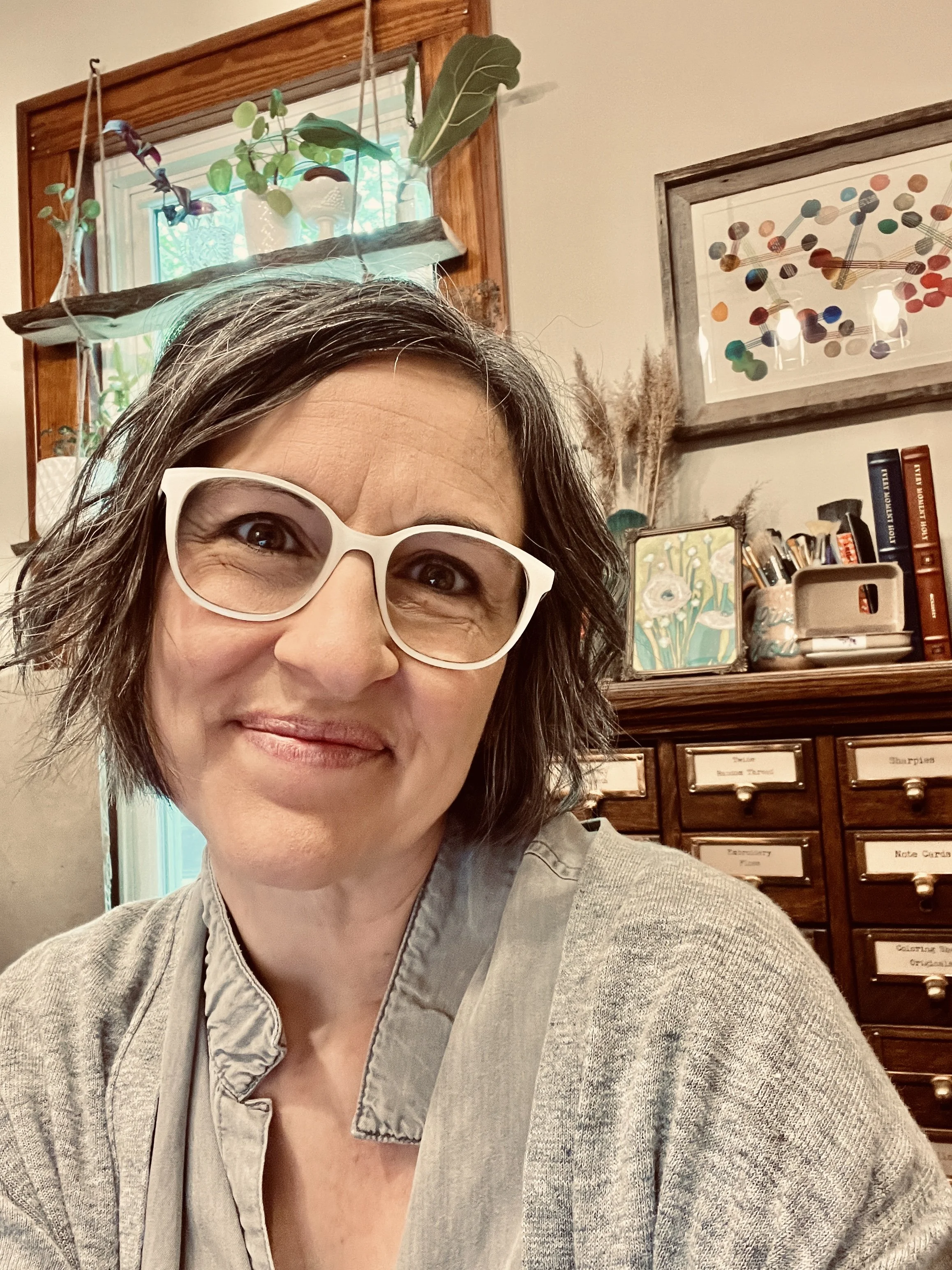 Monica Crandell, an artist with short, wavy dark hair and white glasses smiling at the camera. She's in her cozy art studio with an antique wooden cabinet filled with labeled drawers, artwork, and potted plants on a window shelf behind her.