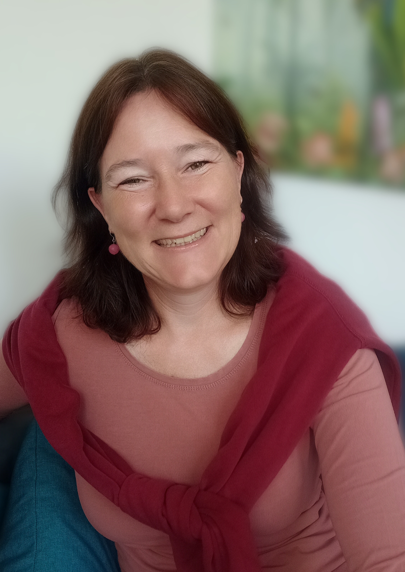 Eine lachende Frau mit dunklen haaren in einem rosa Oberteil mit einem roten Pulli über den Schultern.