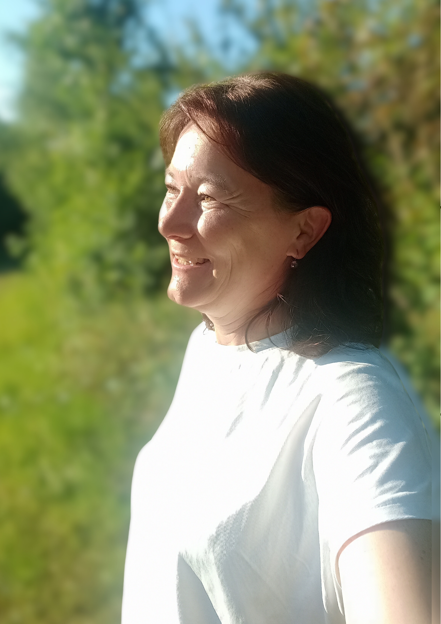 Nahaufnahme eines lächelnden Frau im Freien bei Sonnenlicht, mit grünen Bäumen im Hintergrund.