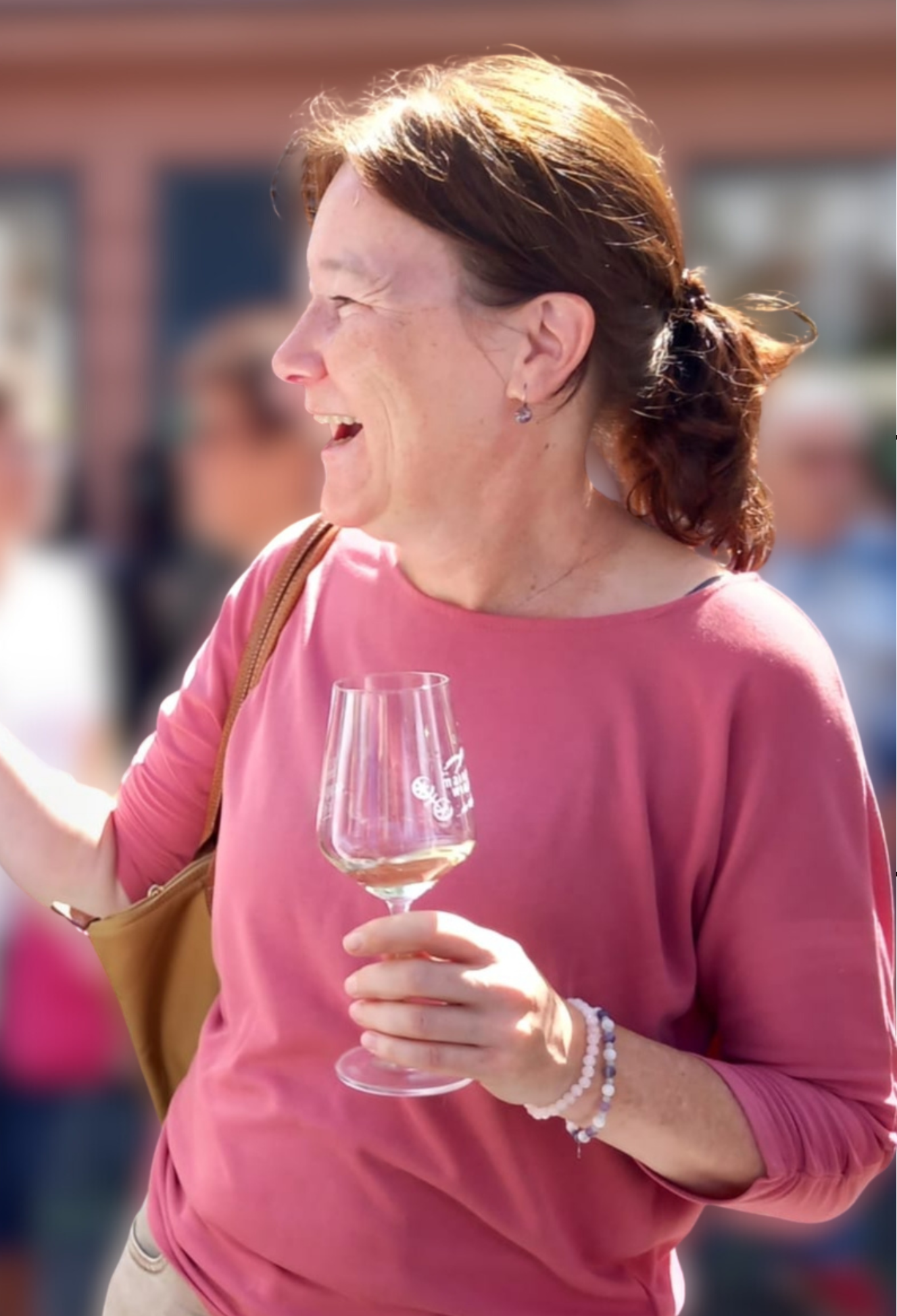 Lächelnde Frau mit braunen Haaren in einem rosa Oberteil, die ein Weinglas hält, vermutlich bei einem Weinfest oder hoher gesellschaftlicher Anlass, im Freien bei Tageslicht.
