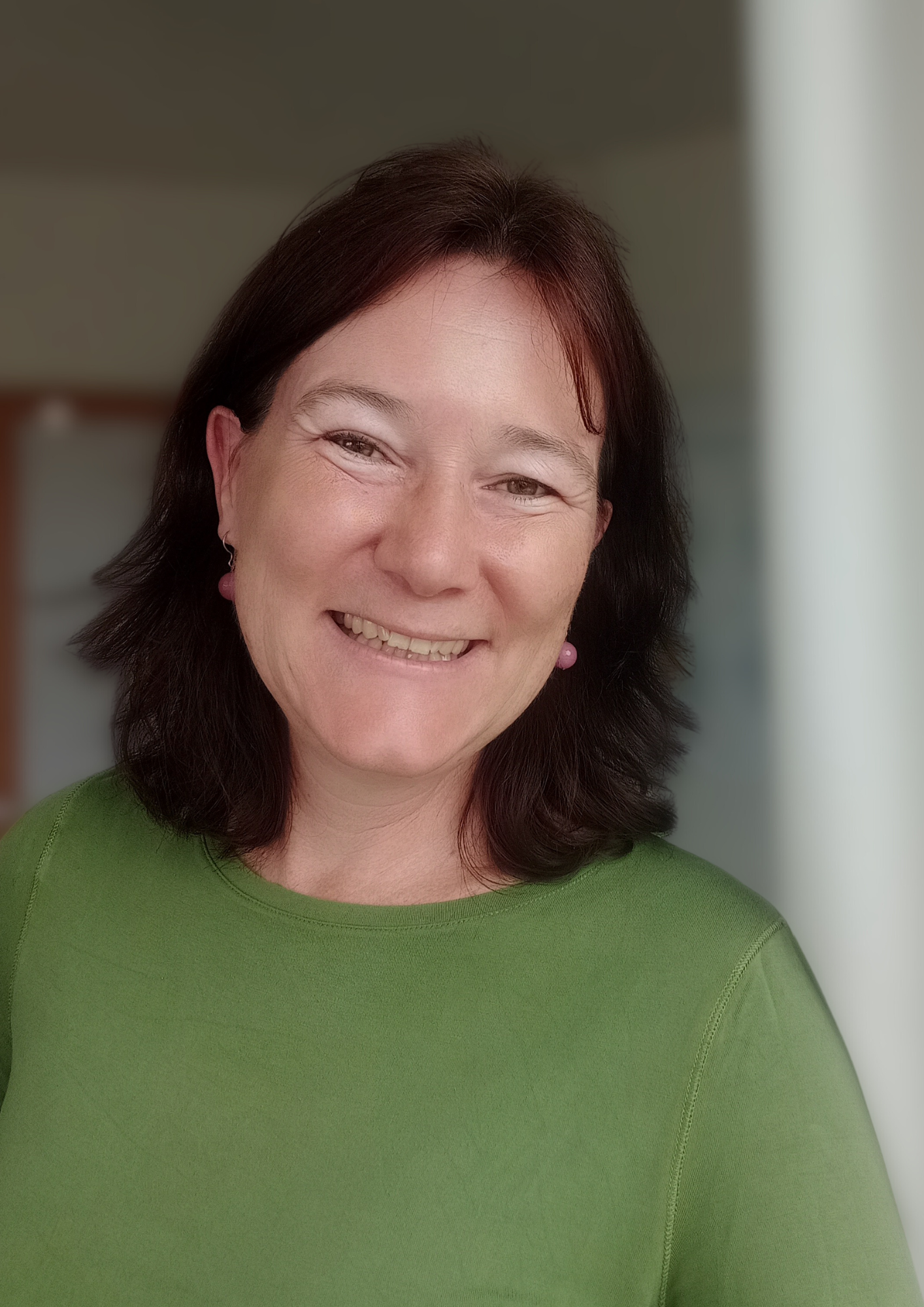 Lächelnde Frau mit dunklem Haar und grünem Pullover, im Hintergrund unscharf, helles Licht.