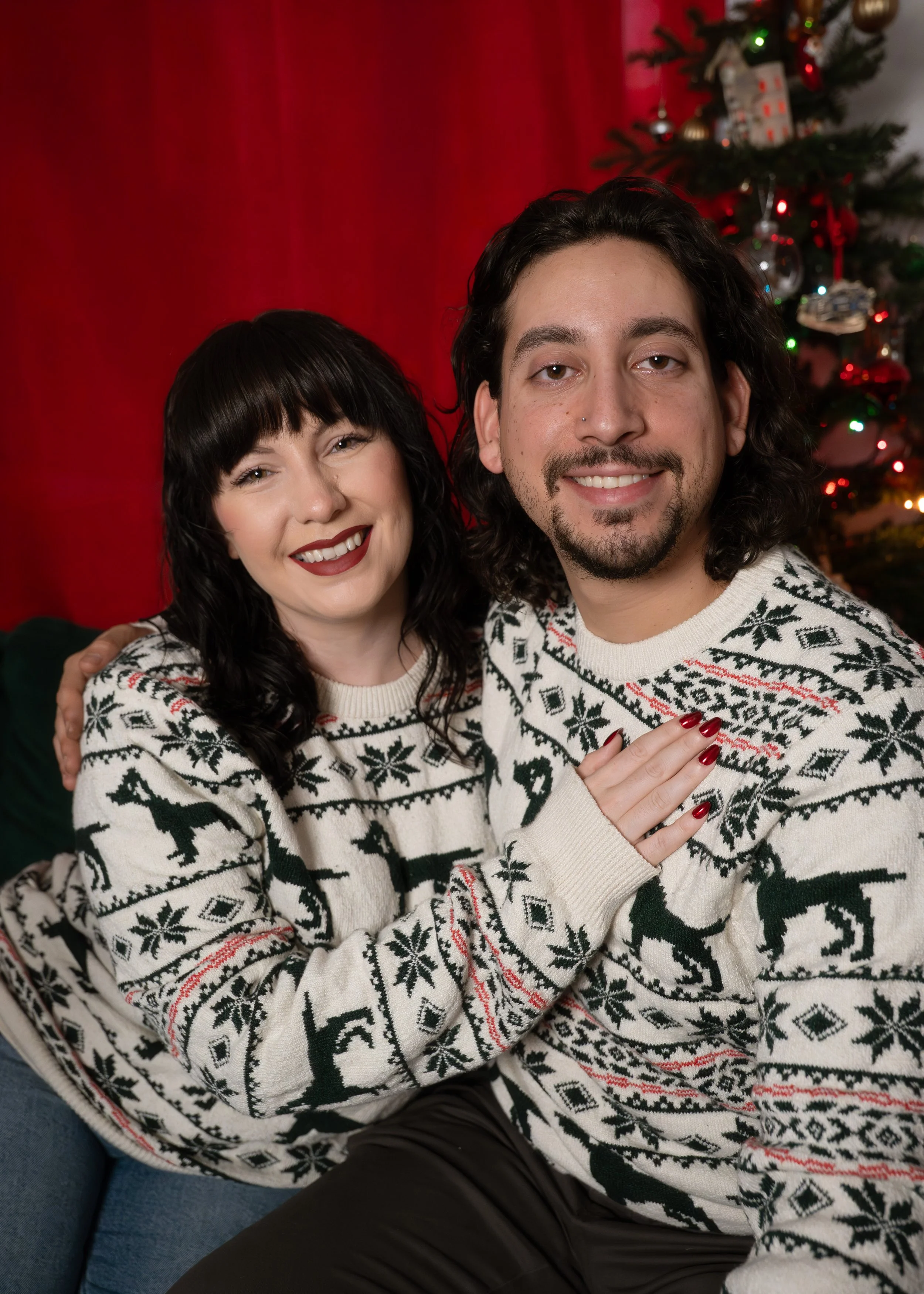 Image of a couple, man and woman, embracing in front of the Christmas tree while wearing holiday sweaters.