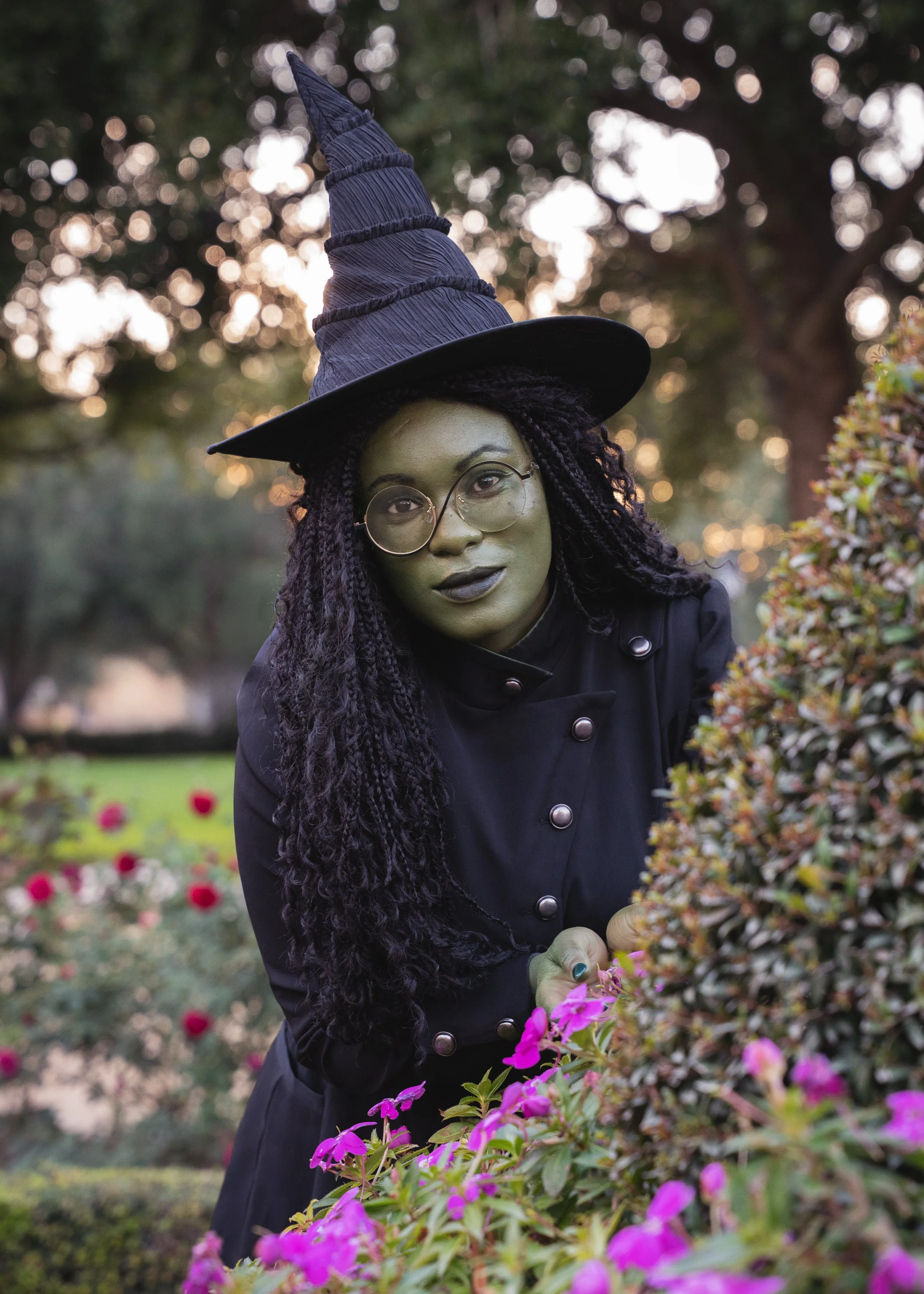 Image of woman dressed as Elphaba, leaning over a flower bush smiling softly.