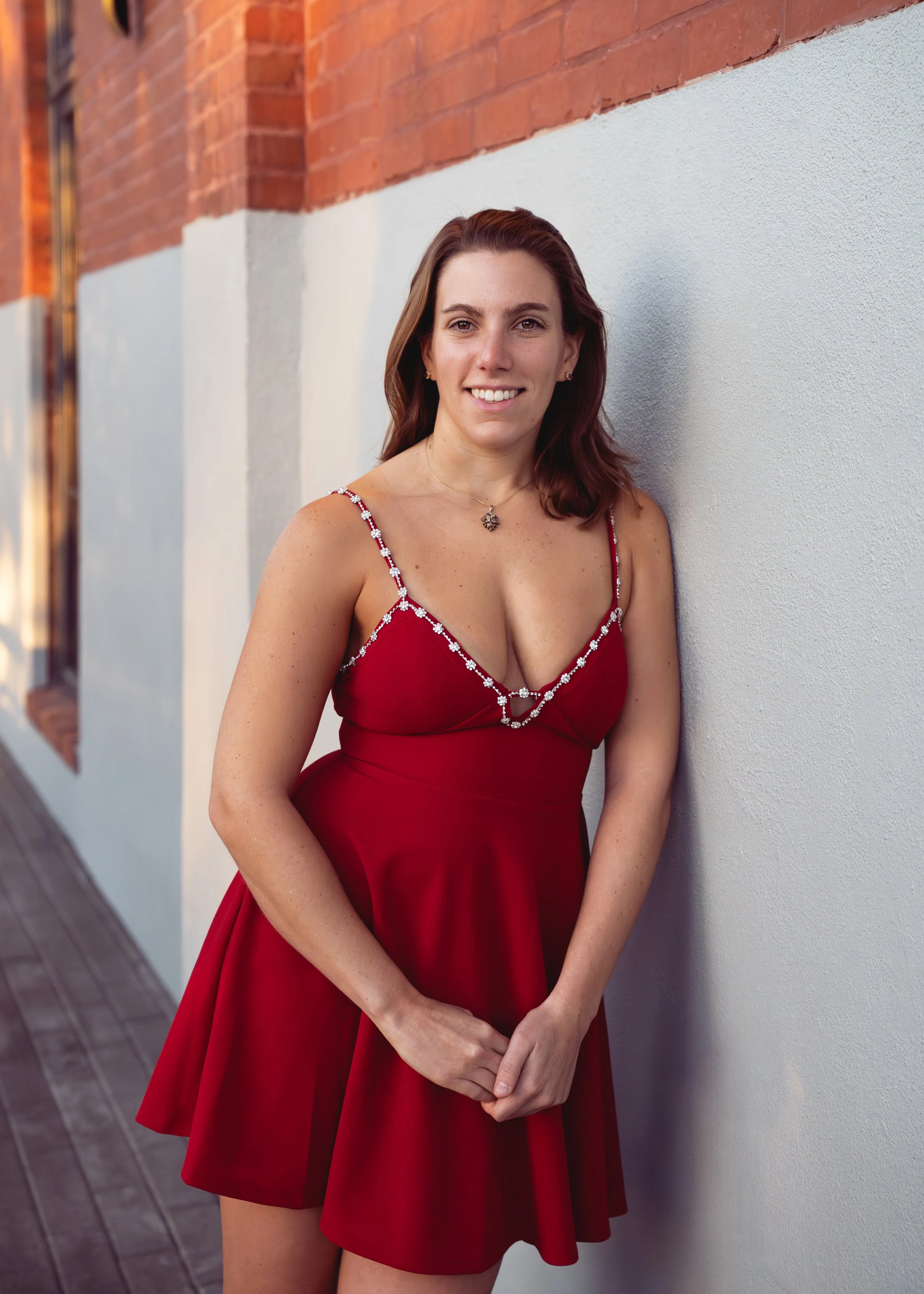 Image of woman wearing red cocktail dress, posing against a wall with a soft smile.